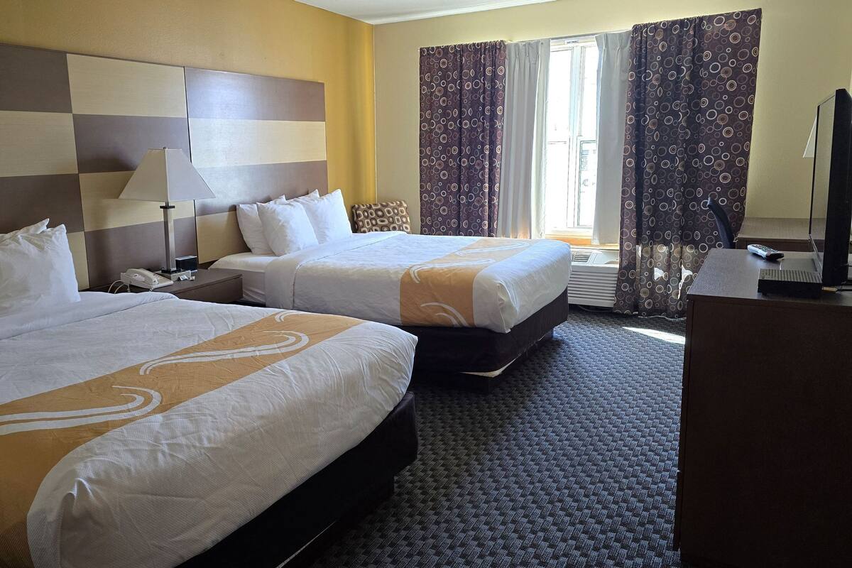 A hotel room with two beds, a desk, a lamp, and a TV.