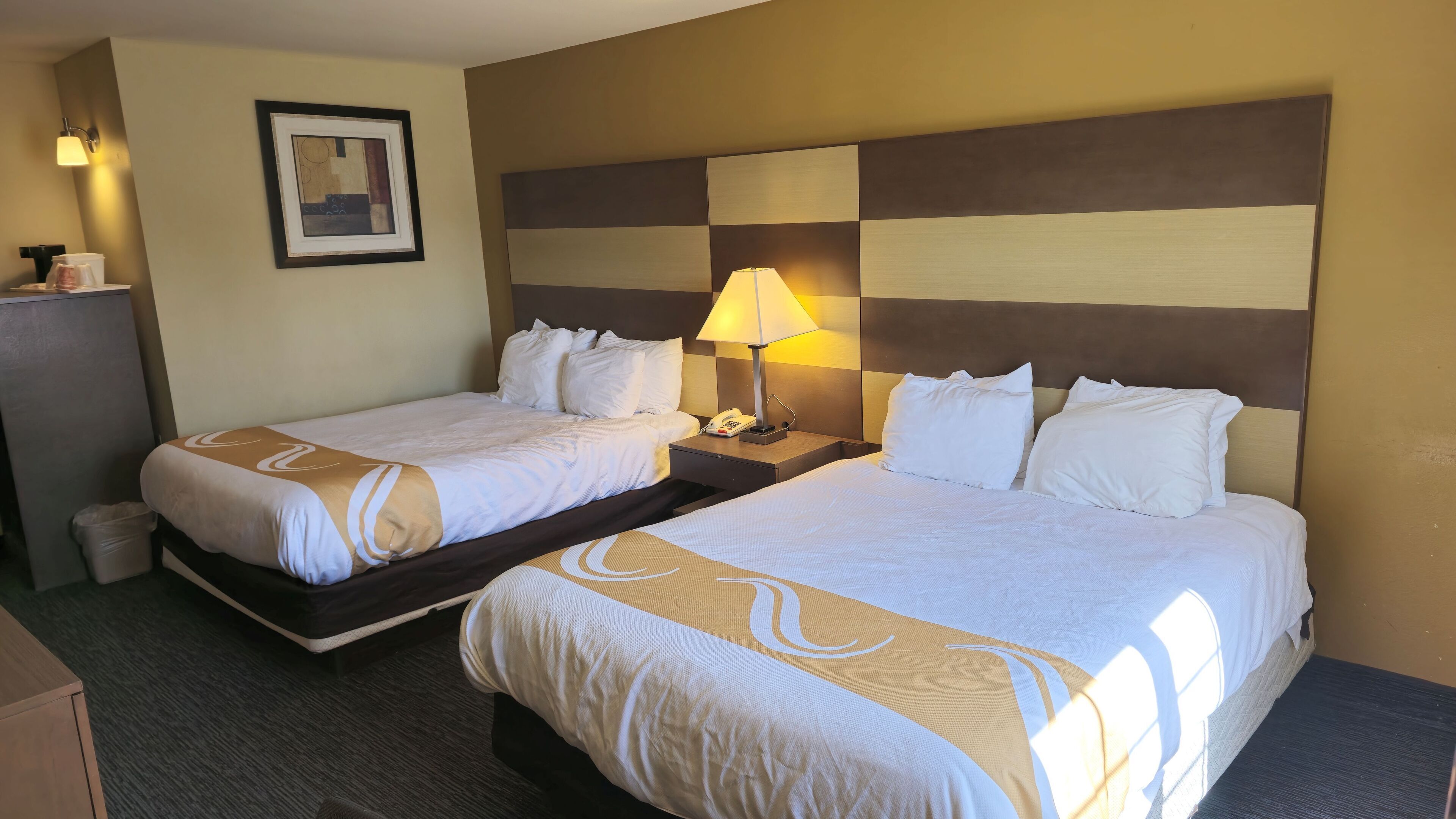 Quarto de hotel com duas camas, mesa de cabeceira com abajur, quadro na parede e um cesto de lixo.