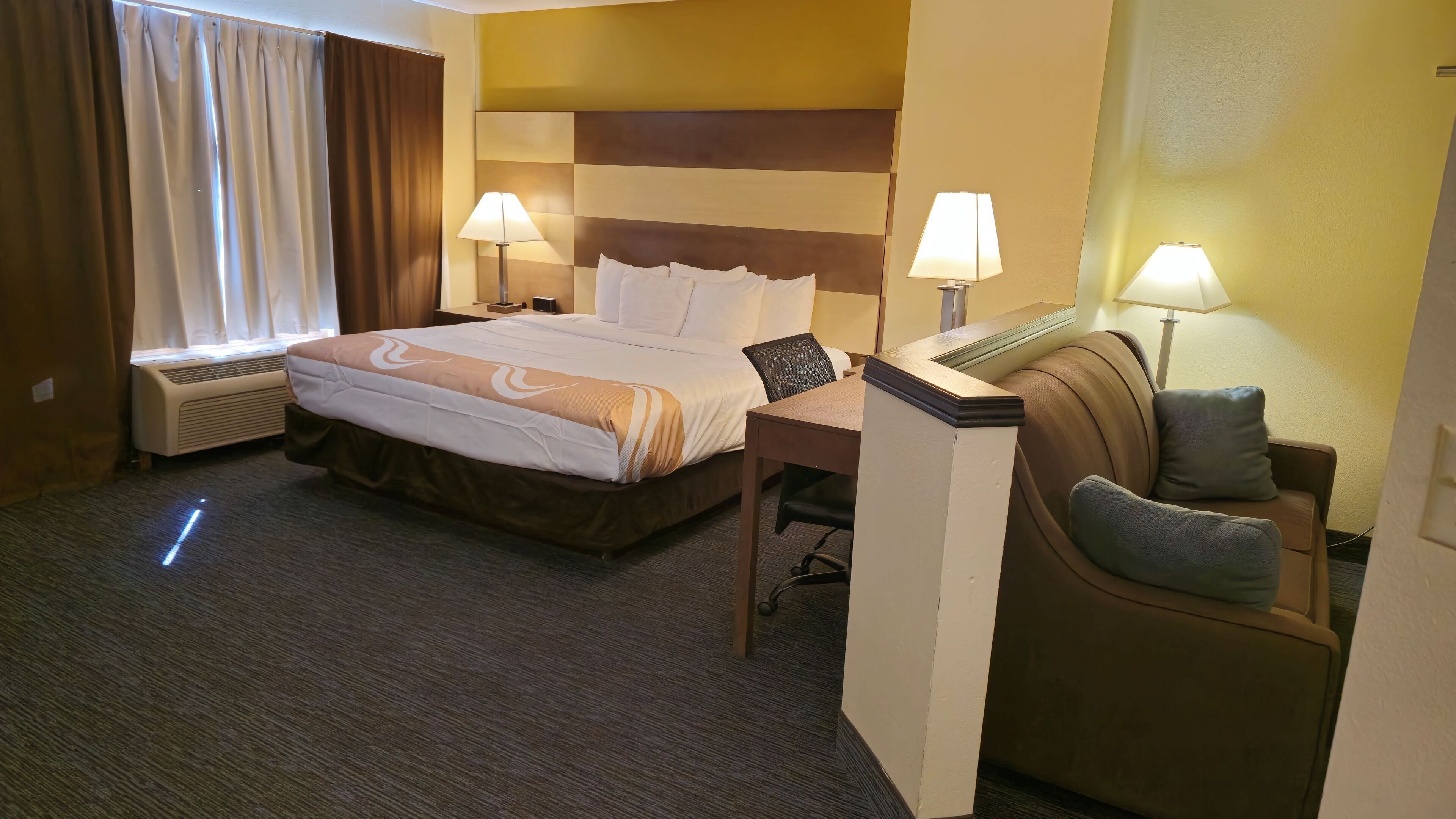 A hotel room with a bed, two bedside lamps, a desk, a chair, and a window with curtains.