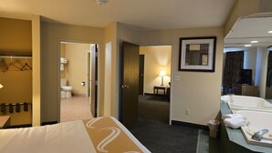 A hotel room with a bed, a bathroom with a bathtub, a mirror, and a hallway leading to another room.