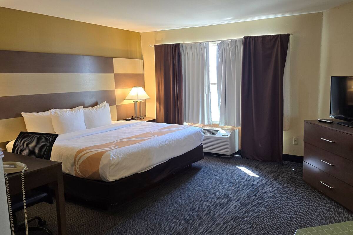 A hotel room with a large bed, a desk with a telephone, a television on a dresser, and a window with curtains.