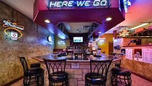 A bar with a neon sign reading "HERE WE GO", a Budweiser sign, and a neon sign for "SHOCK TOP". There are four stools and a bar counter with bottles and glasses.