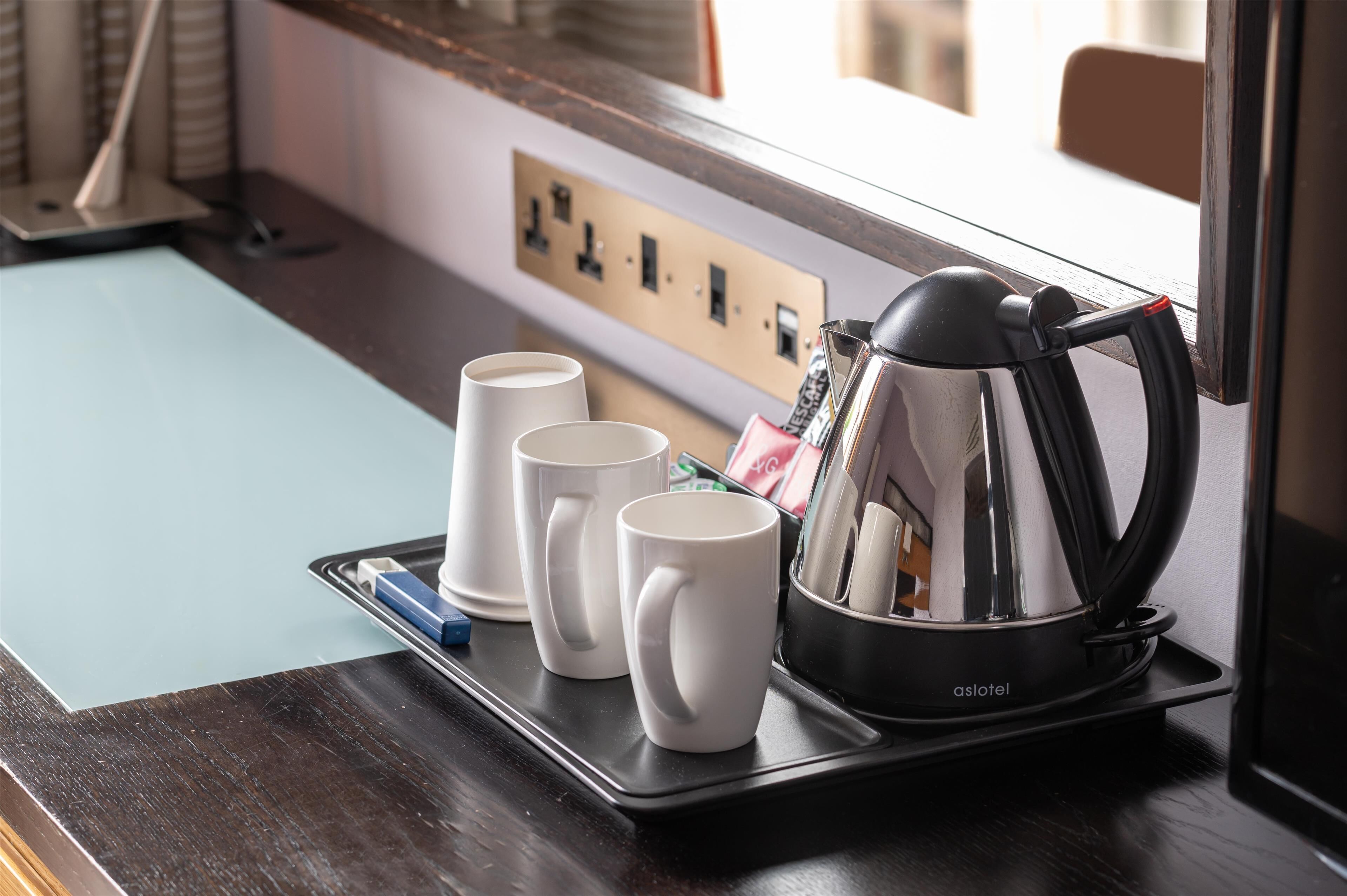 A tray with three white mugs and a black electric kettle on a dark wooden counter near a window.
