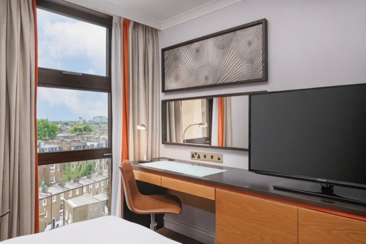 A hotel room with a large window, a desk with a mirror, a television, and a bed.