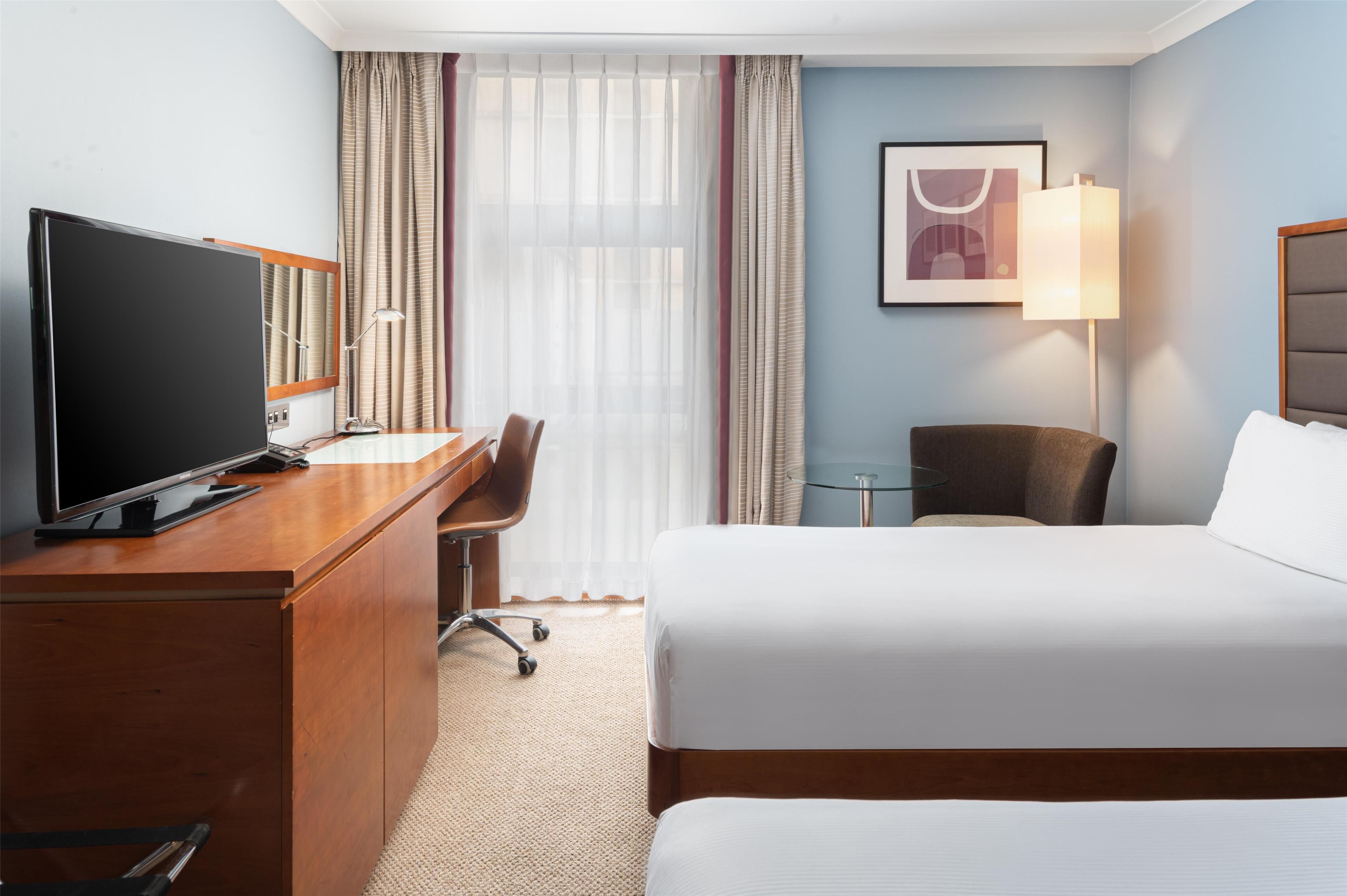 A hotel room with a bed, a desk with a TV, a chair, a lamp, and a framed picture on the wall.