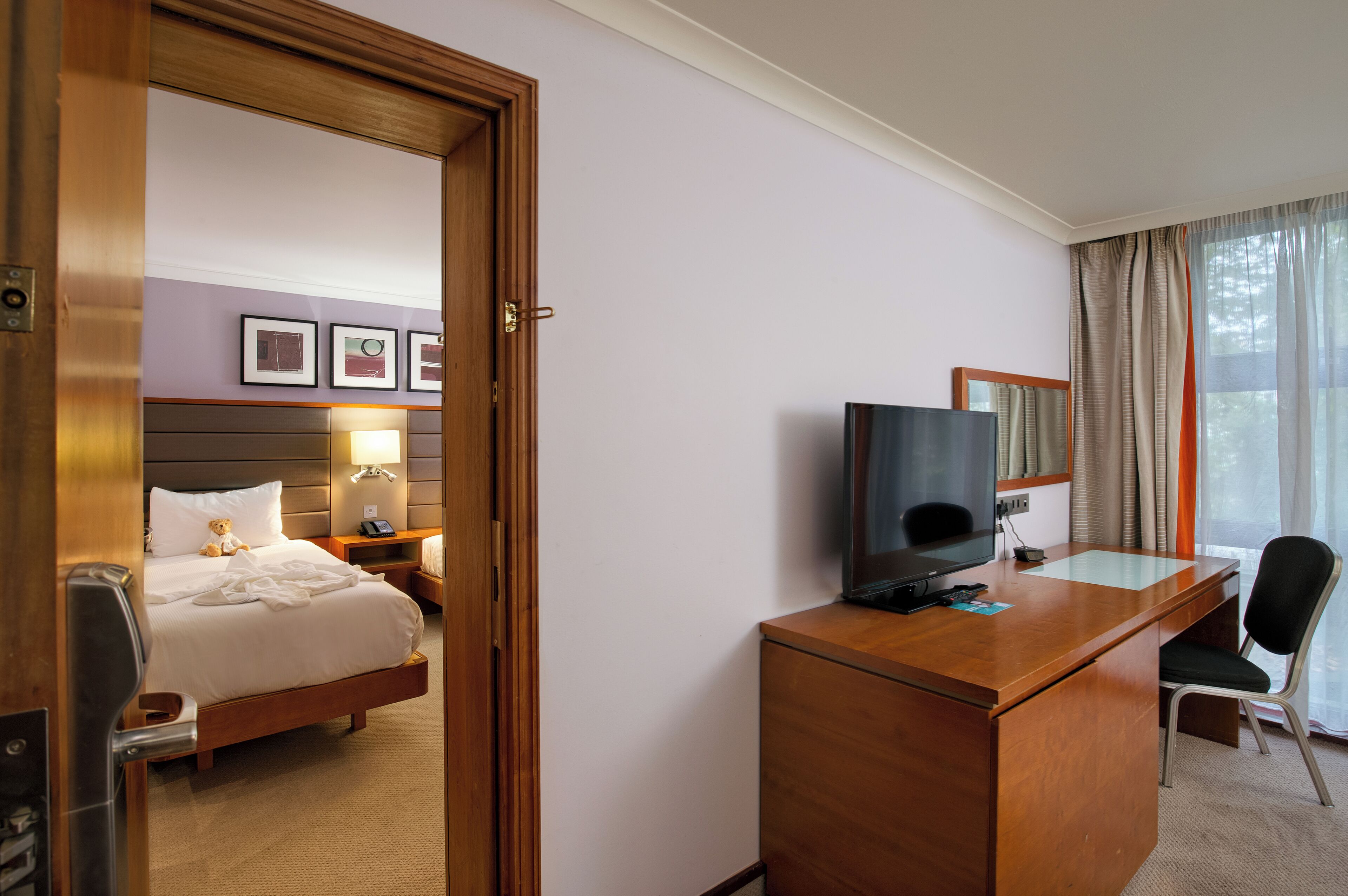 Ein Hotelzimmer mit einem Bett, einem Schreibtisch mit Fernseher, einem Sessel und einem Fenster mit Vorhängen.