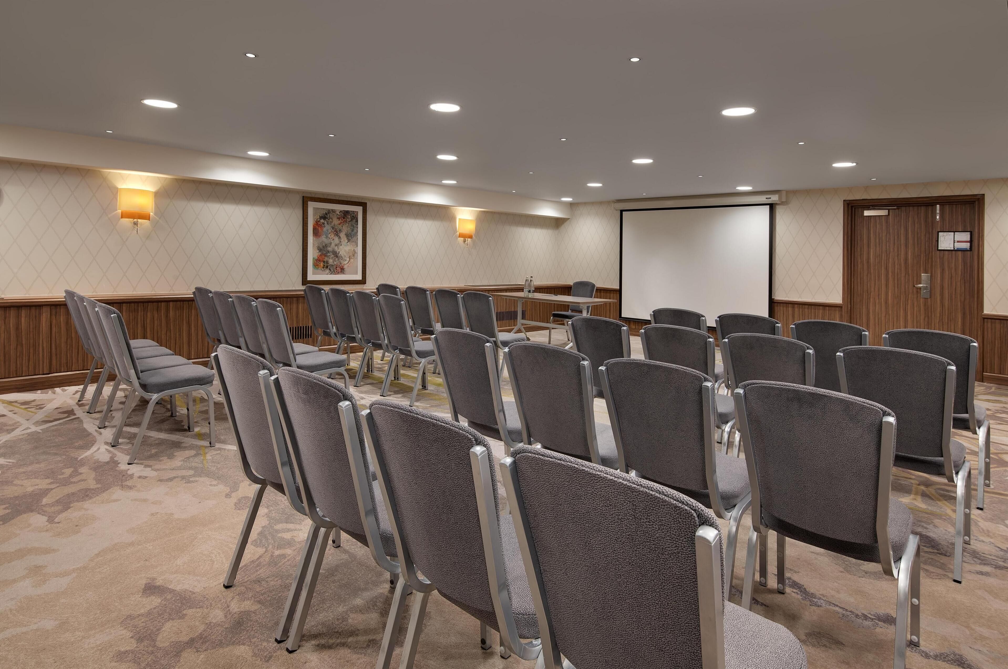 A conference room with rows of grey chairs, a projector screen, and a wooden panel wall.