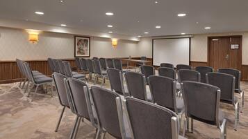 A conference room with rows of grey chairs, a projector screen, and a wooden panel wall.