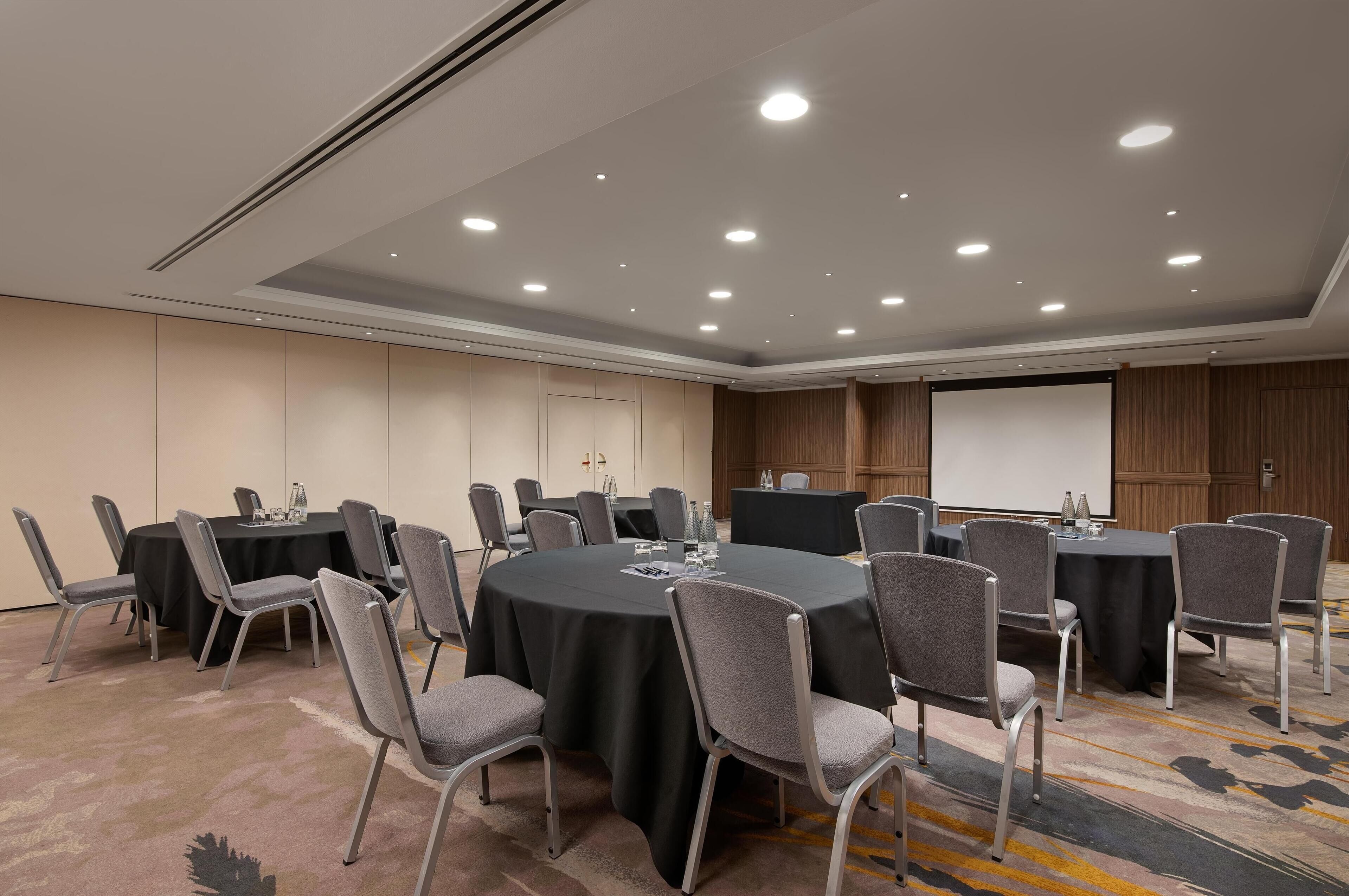 Une salle de conférence avec des tables rondes, des chaises grises et un écran de projection.