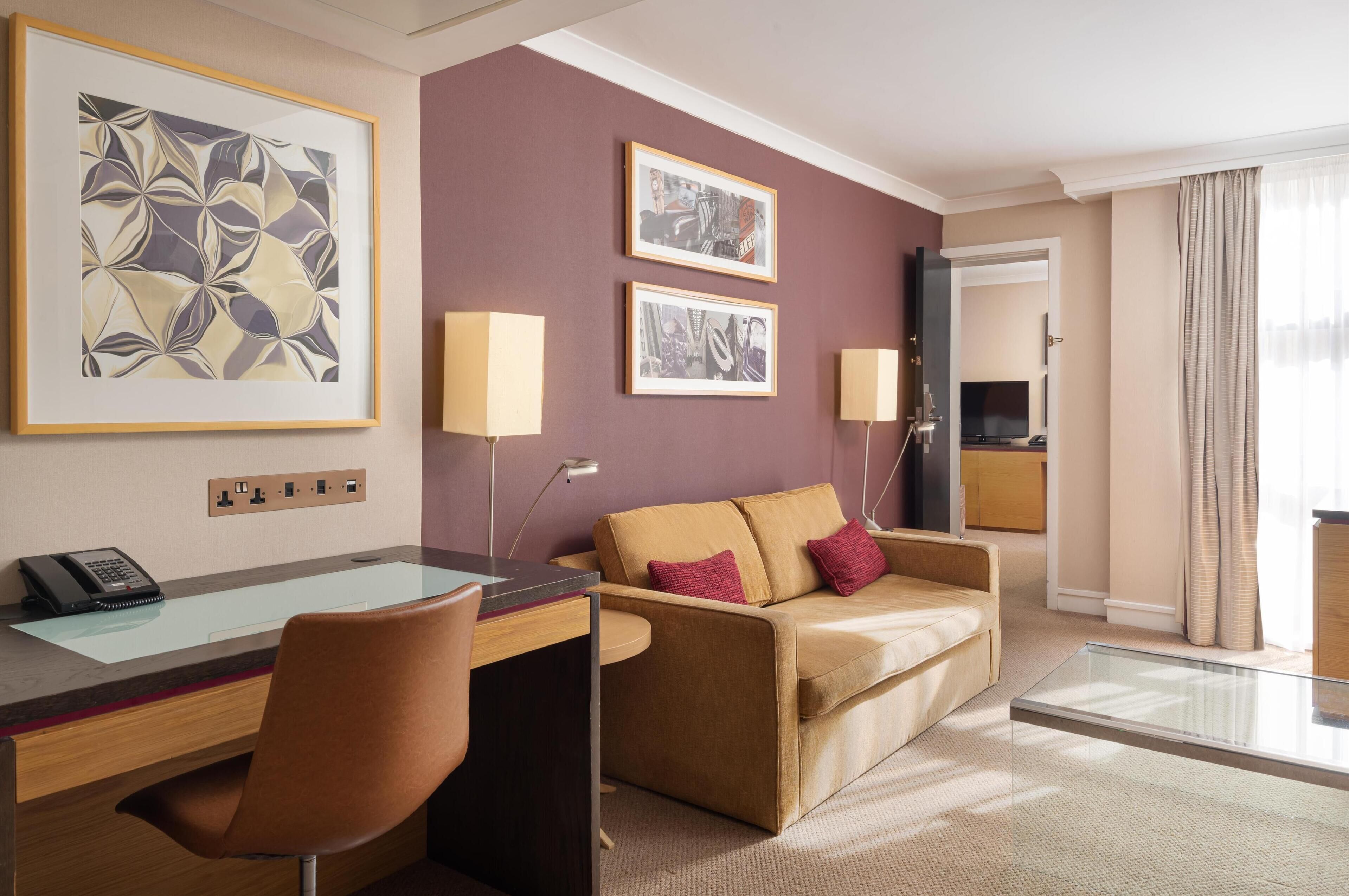 Ett hotellrum med ett skrivbord, en stol, en soffa och två inramade tavlor på väggen.
