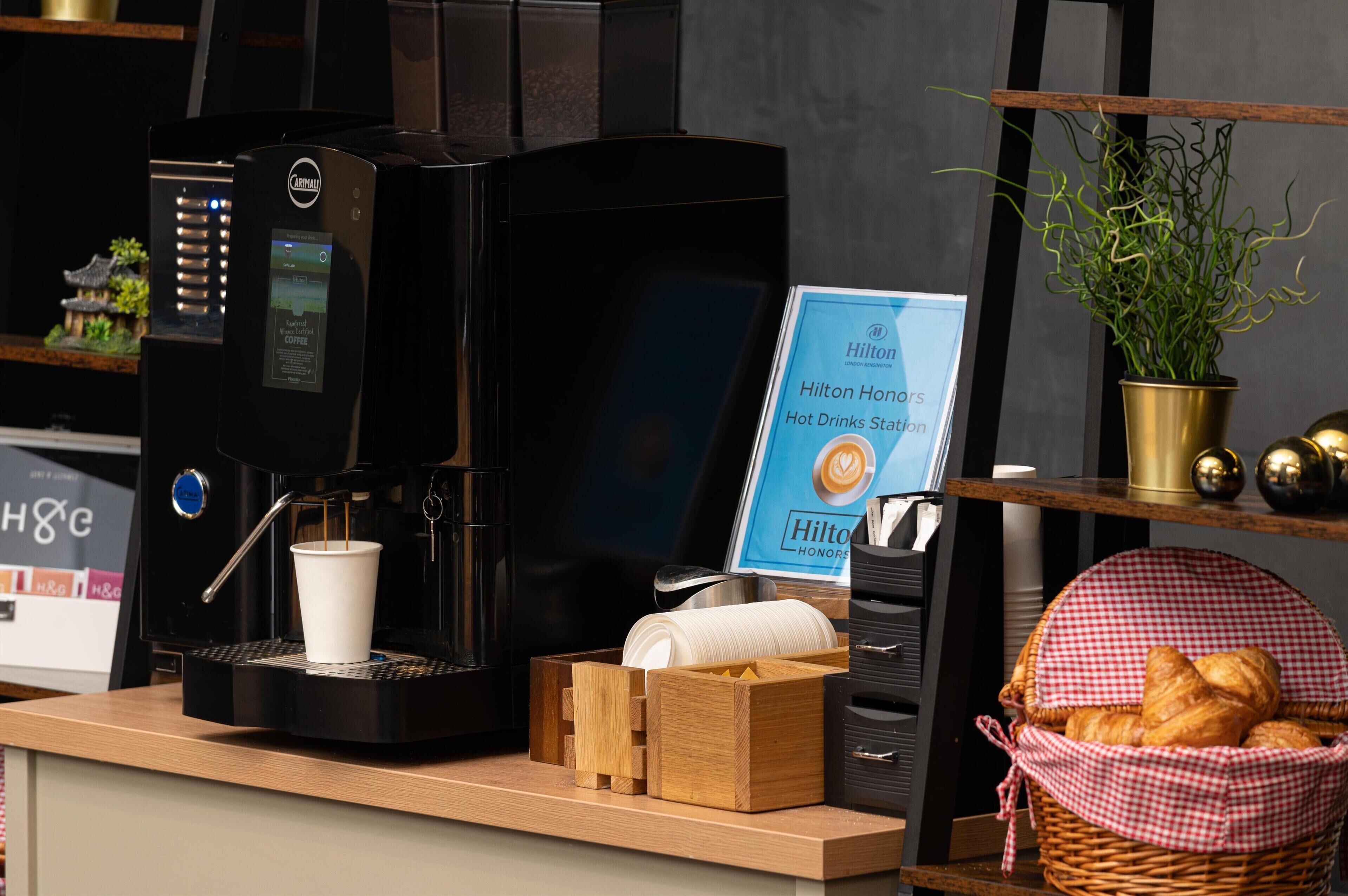 Una máquina de café, un letrero que indica Estación de Bebidas Calientes Hilton Honors, una cesta de cruasanes y una planta en maceta.