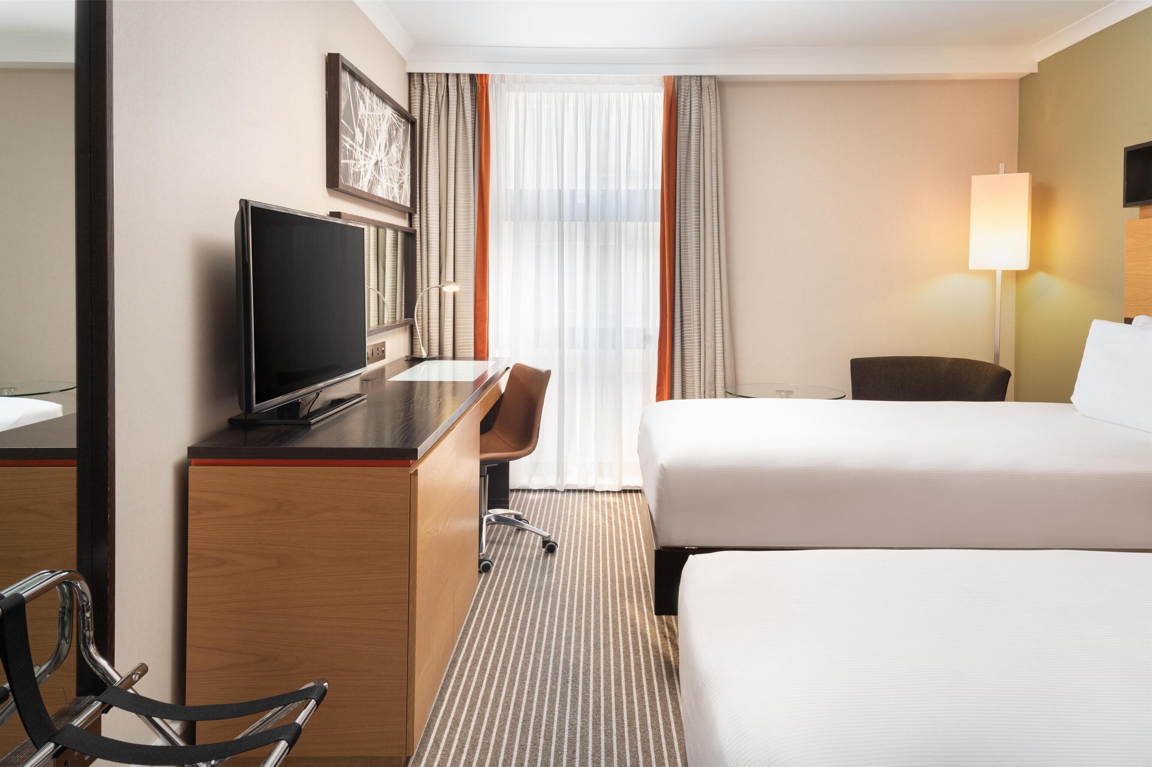 A hotel room with a bed, a desk with a television, a chair, a lamp, and a window with curtains.