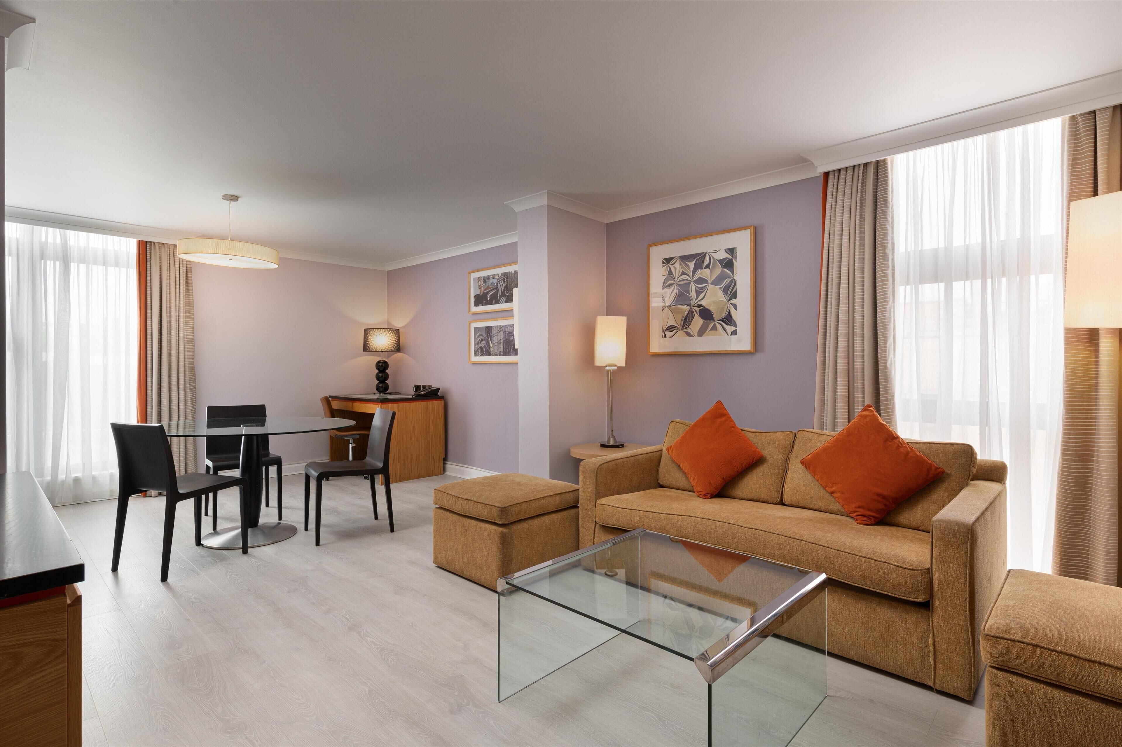 A modern living room with a glass coffee table, a beige sofa with orange cushions, a dining area with a table and chairs, and artwork on the walls.