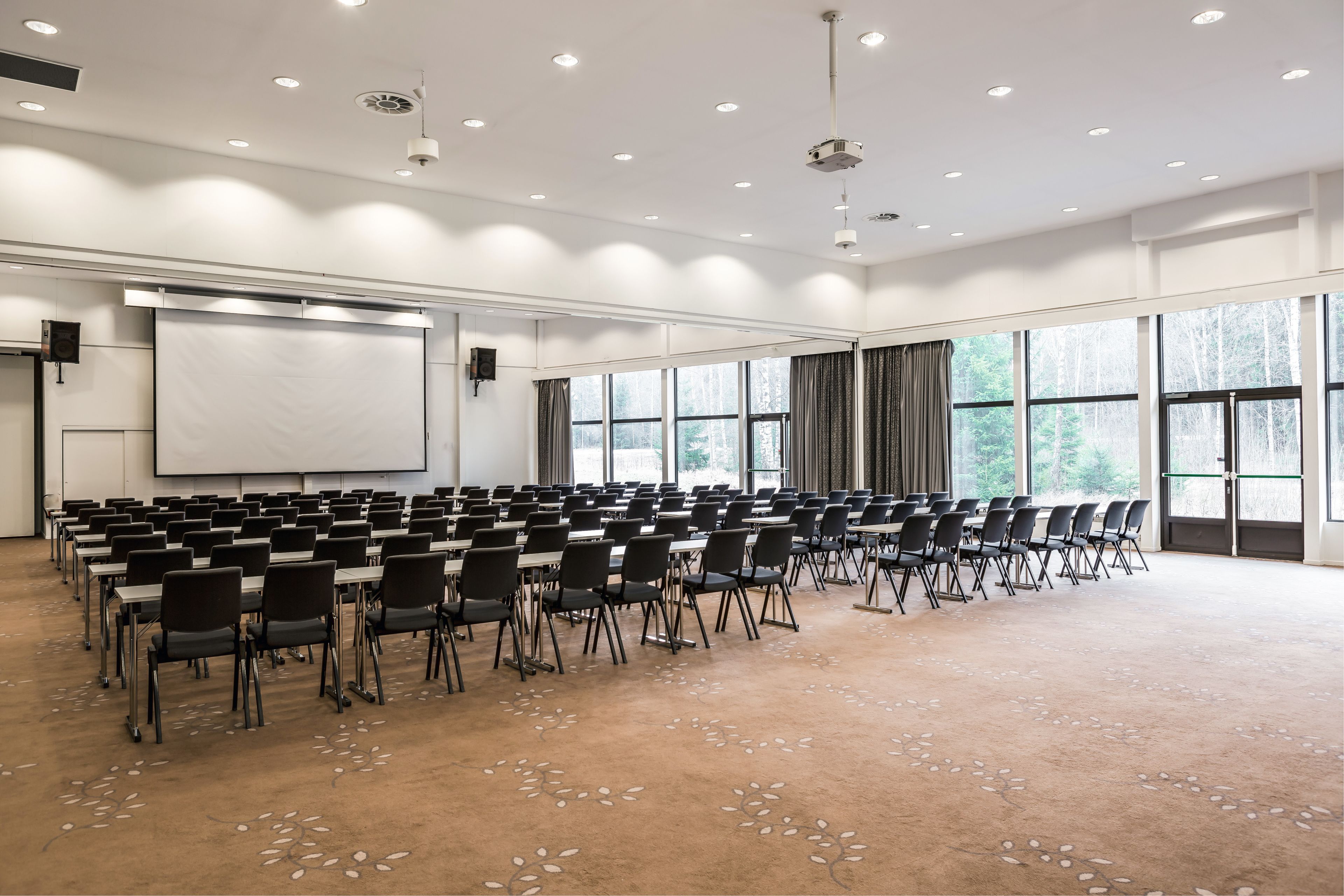 A large conference room with rows of tables and chairs, a projector screen, and large windows.