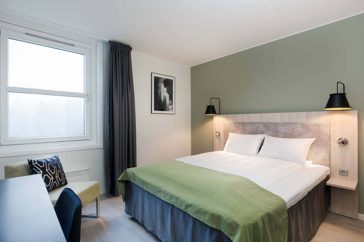 A hotel room with a bed, bedside tables, a chair, a window with curtains, and a wall-mounted lamp.