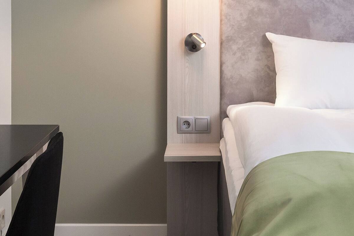 A neatly made bed with white linens and a green throw, a bedside table with a lamp, and a wall socket.