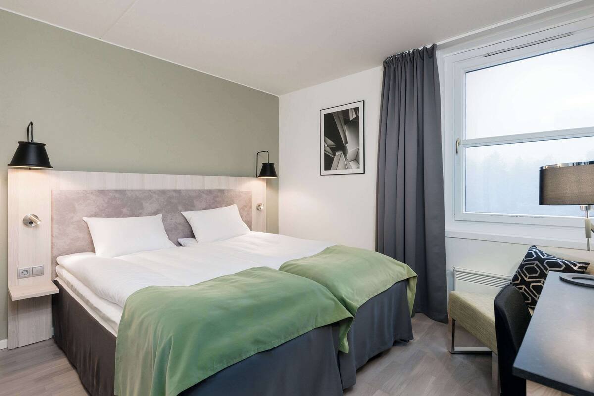 A hotel room with a bed, bedside lamps, a window with curtains, a desk, and a chair.