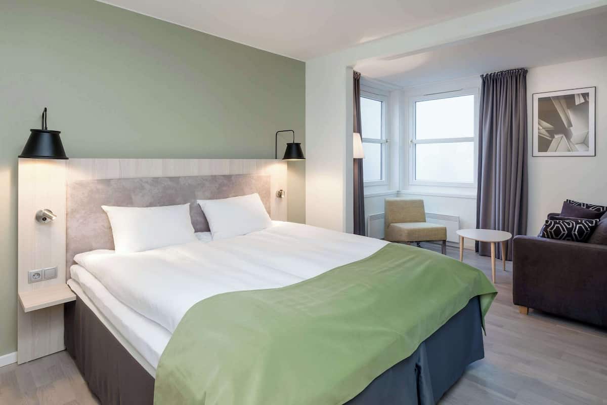 A hotel room with a large bed, bedside tables, a sofa, a small table, and a window with curtains.