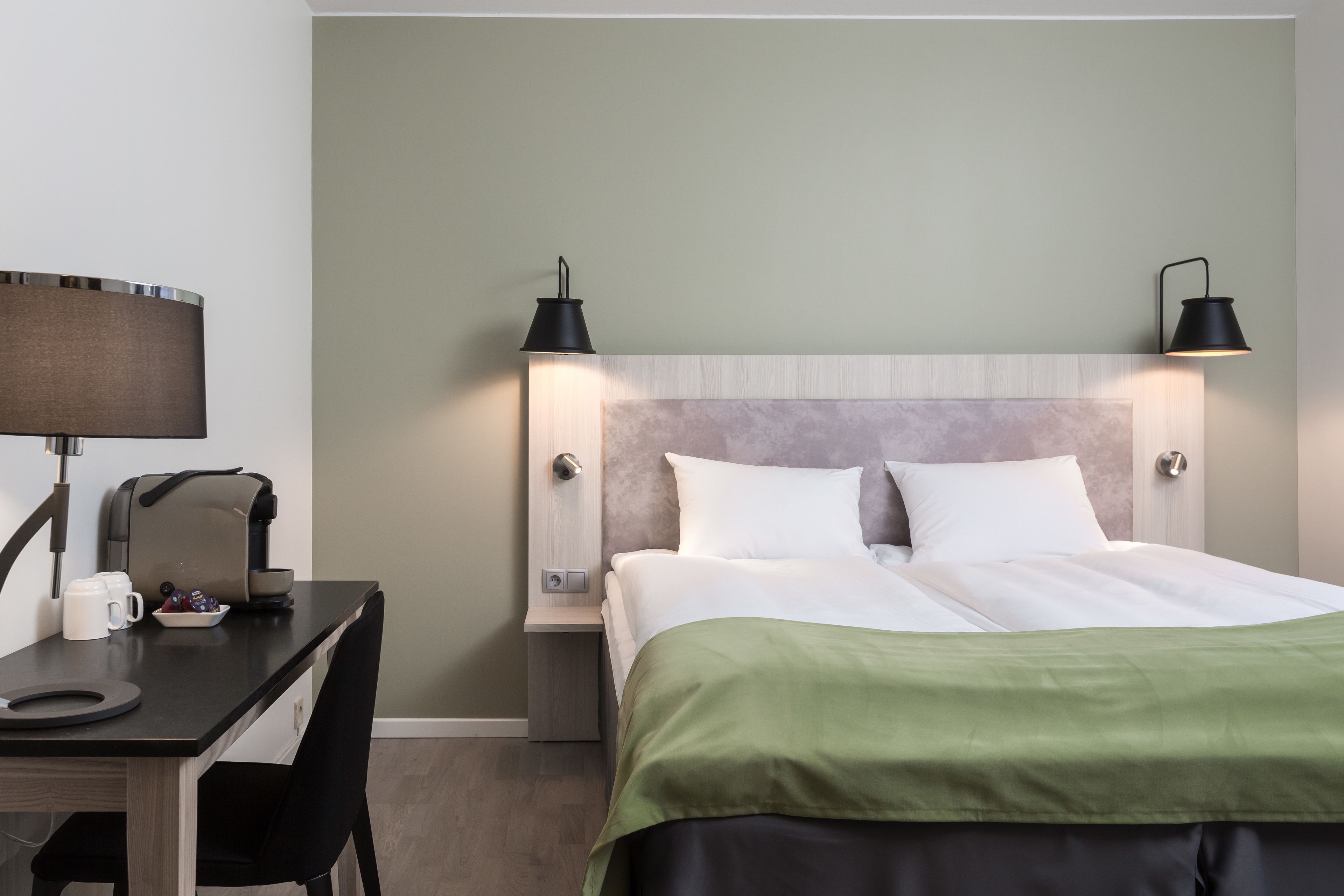 Una habitación de hotel con cama, escritorio con lámpara, cafetera y dos lámparas de noche.