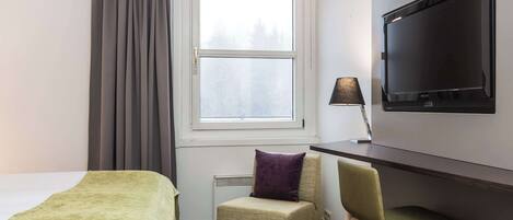 A hotel room with a bed, a chair, a desk with a television, and a window with curtains.