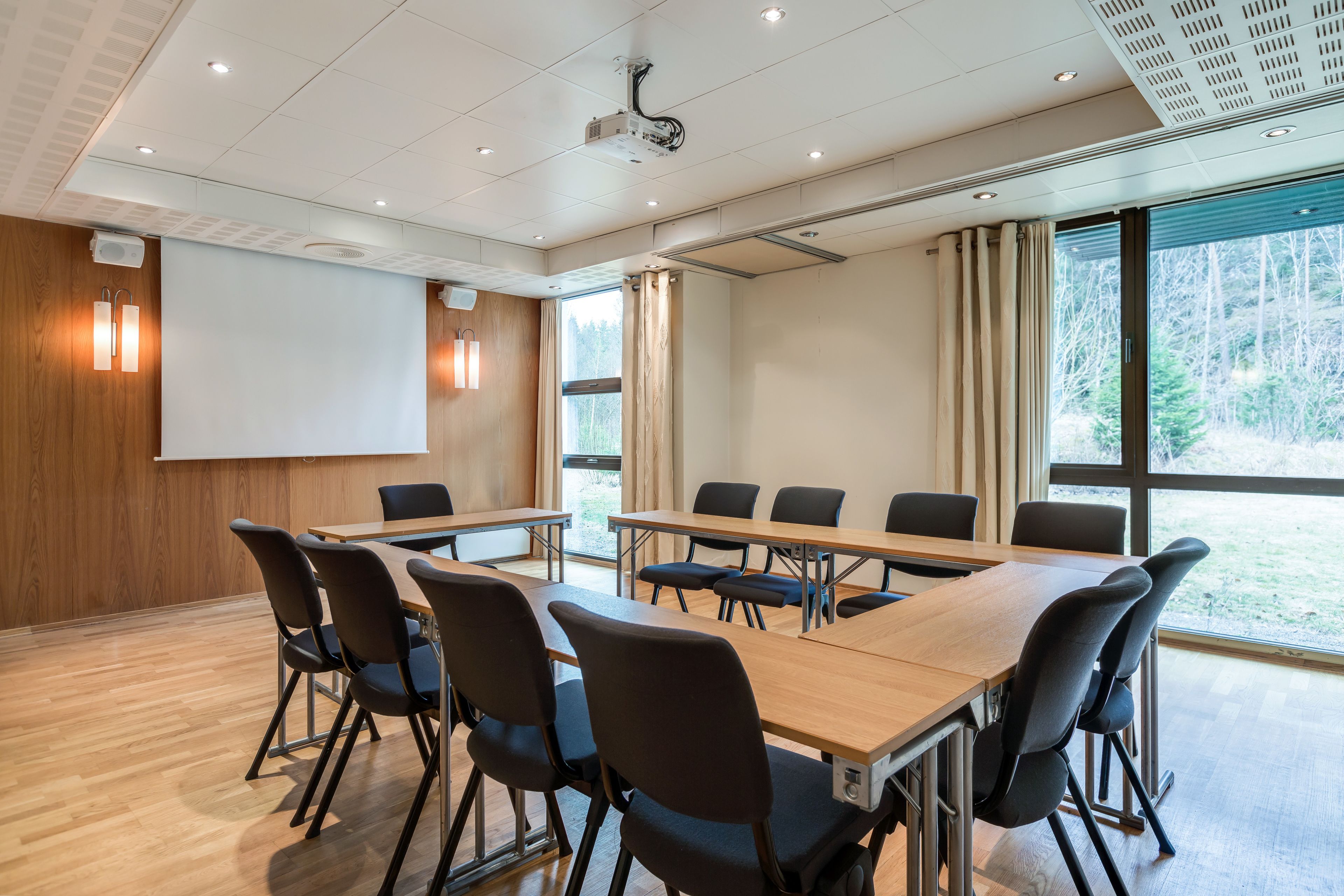A conference room with a long table, chairs, a projector screen, and large windows with curtains.