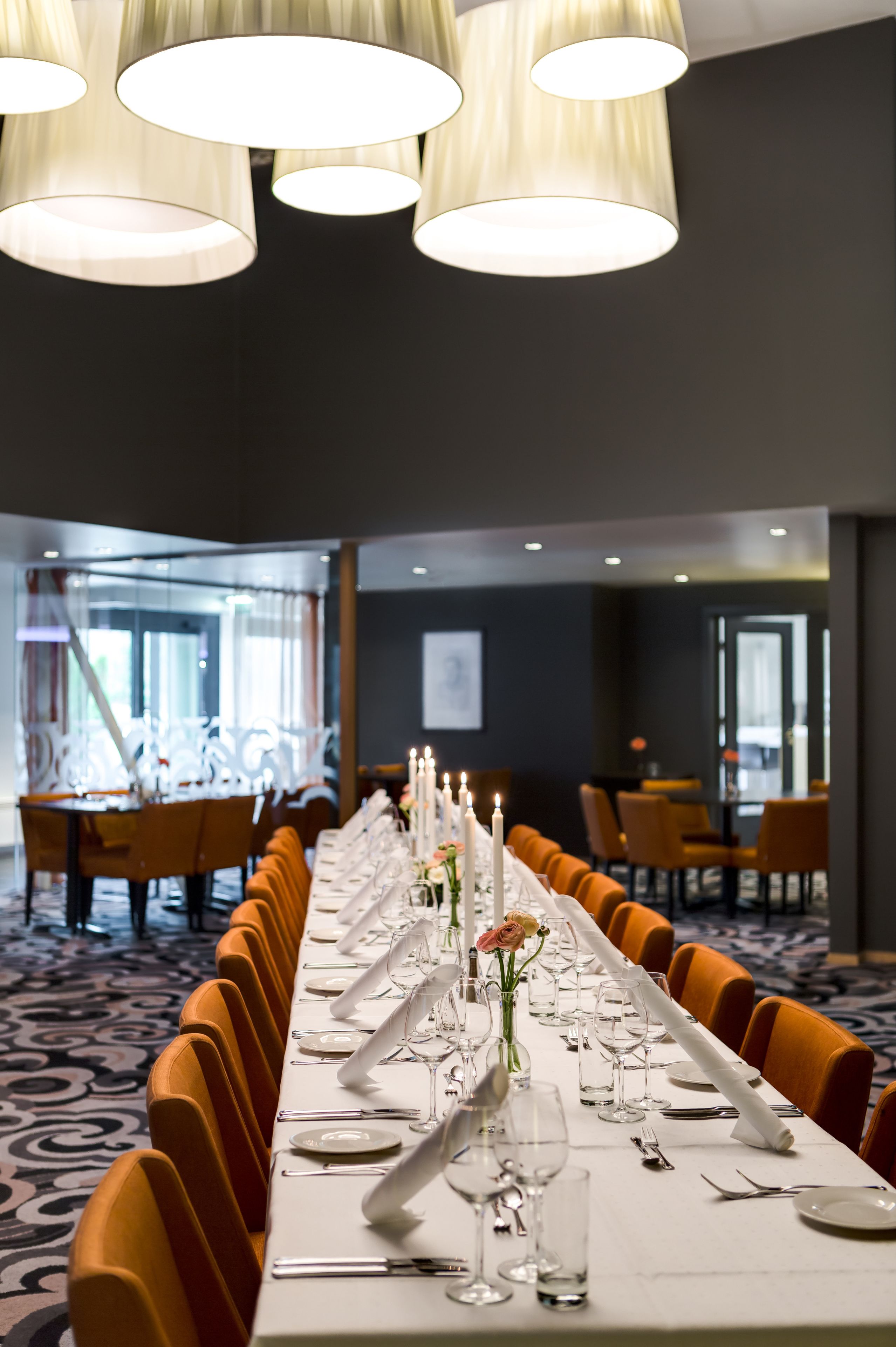 Una larga mesa de comedor con manteles blancos, velas y sillas anaranjadas en una habitación de hotel moderna.