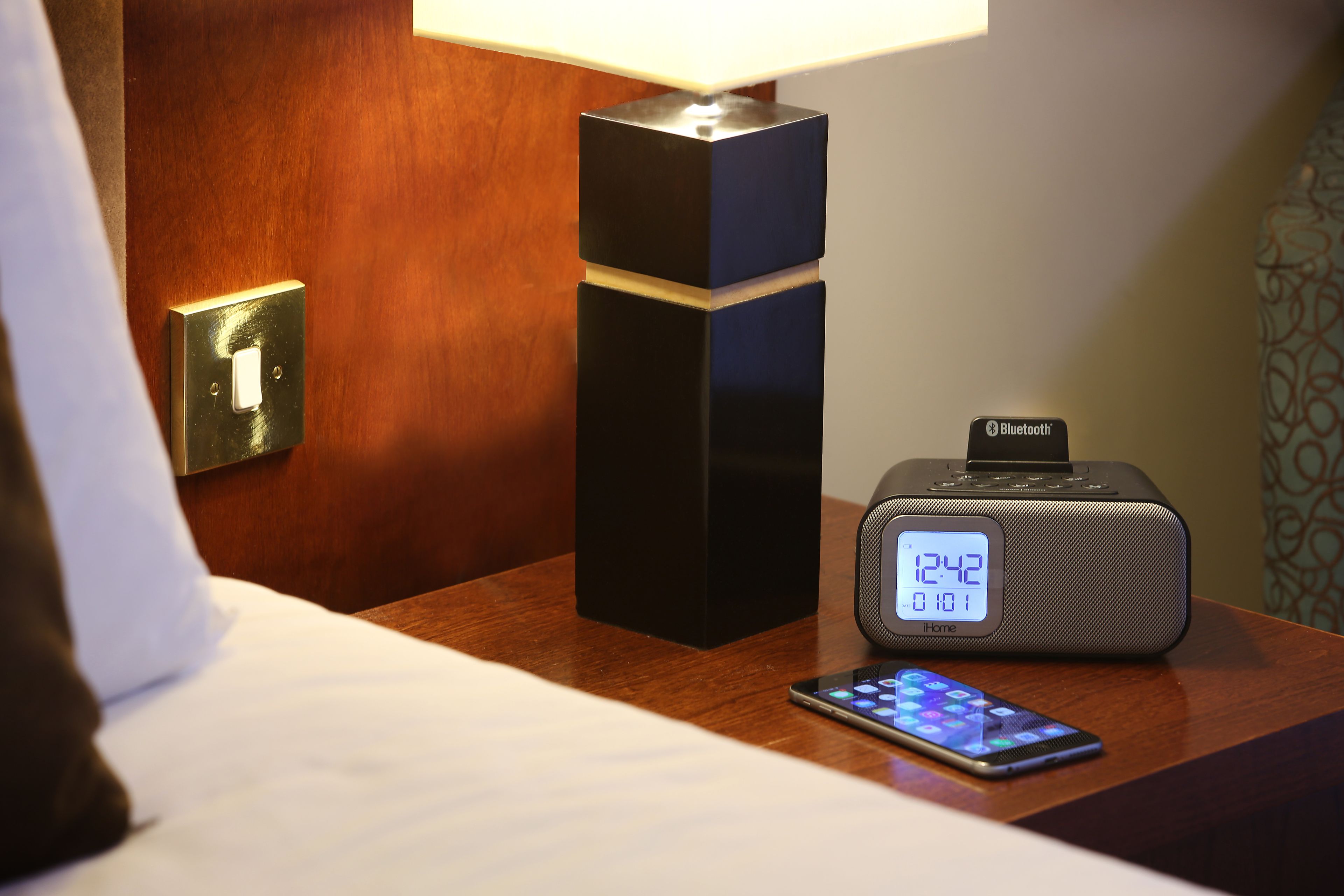 Ein Hotelzimmer mit einem Nachttisch, der eine digitale Uhr, einen Bluetooth-Lautsprecher und ein Smartphone zeigt.