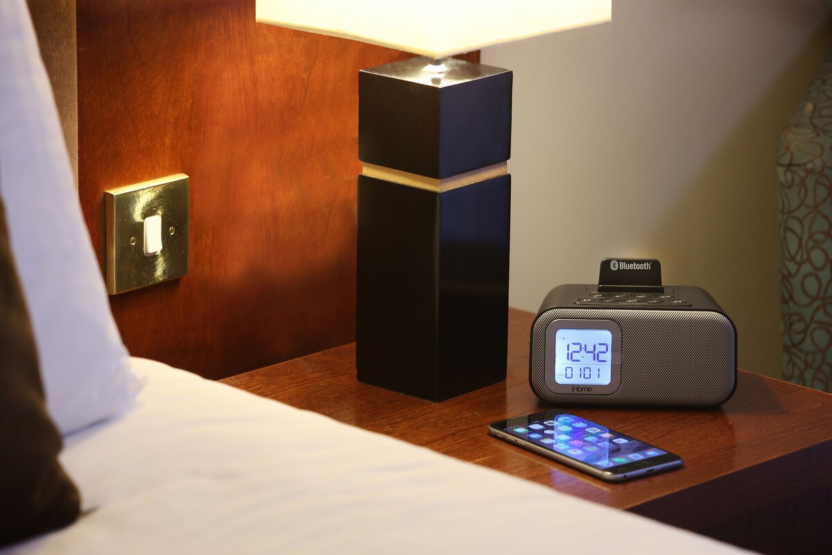 A hotel room with a bedside table featuring a digital clock, a Bluetooth speaker, and a smartphone.