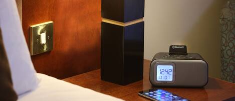 Een hotelkamer met een nachtkastje, waarop een digitale klok, een bluetooth-speaker en een smartphone staan.