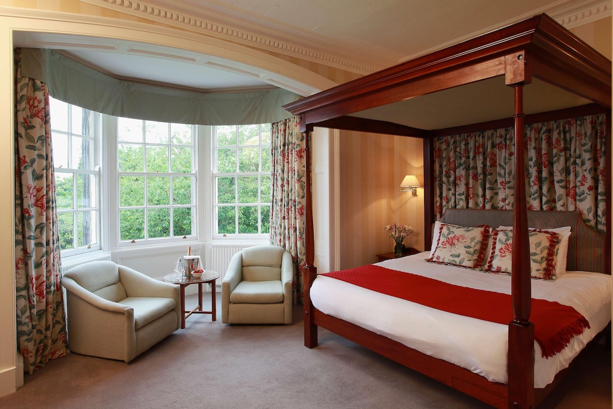 A four-poster bed with floral curtains, a sitting area with two armchairs, and a view of greenery through large windows.
