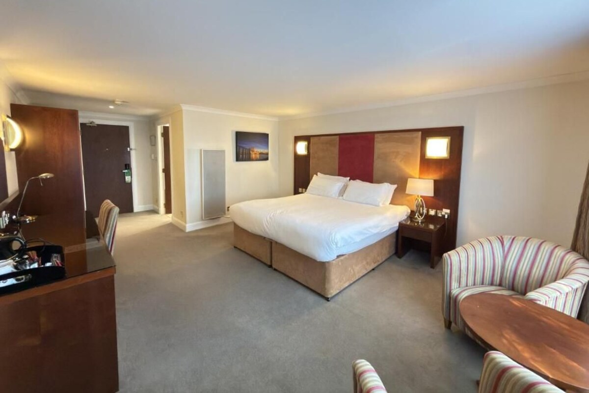 A hotel room with a bed, bedside table, lamp, chair, and a desk.