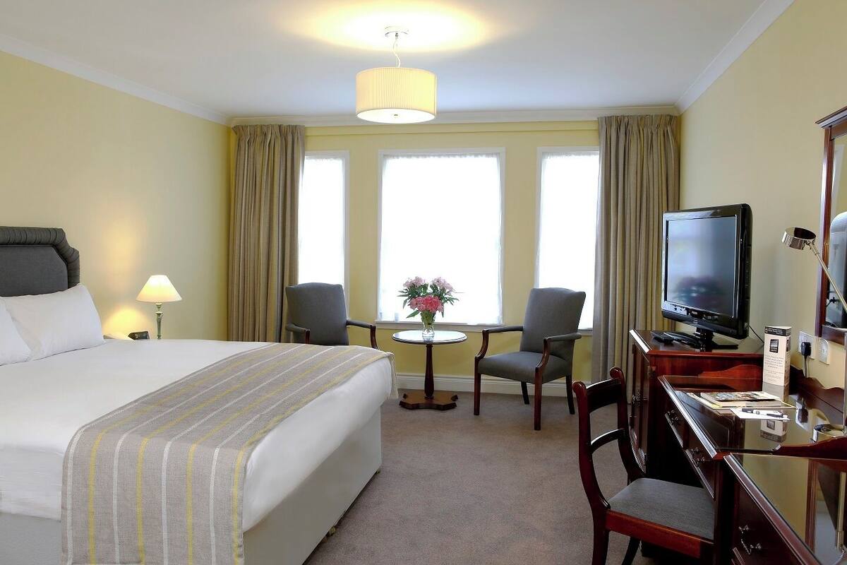 Habitación de hotel con una cama grande, un escritorio con televisor, dos sillas, una mesita redonda con flores y una lámpara.