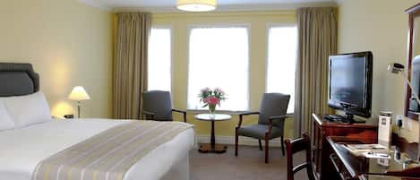 Ett hotellrum med en stor säng, ett skrivbord med en tv, två stolar, ett litet runt bord med blommor och en lampa.