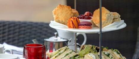 A three-tiered serving tray with sandwiches, scones, and pastries on a glass table outdoors.