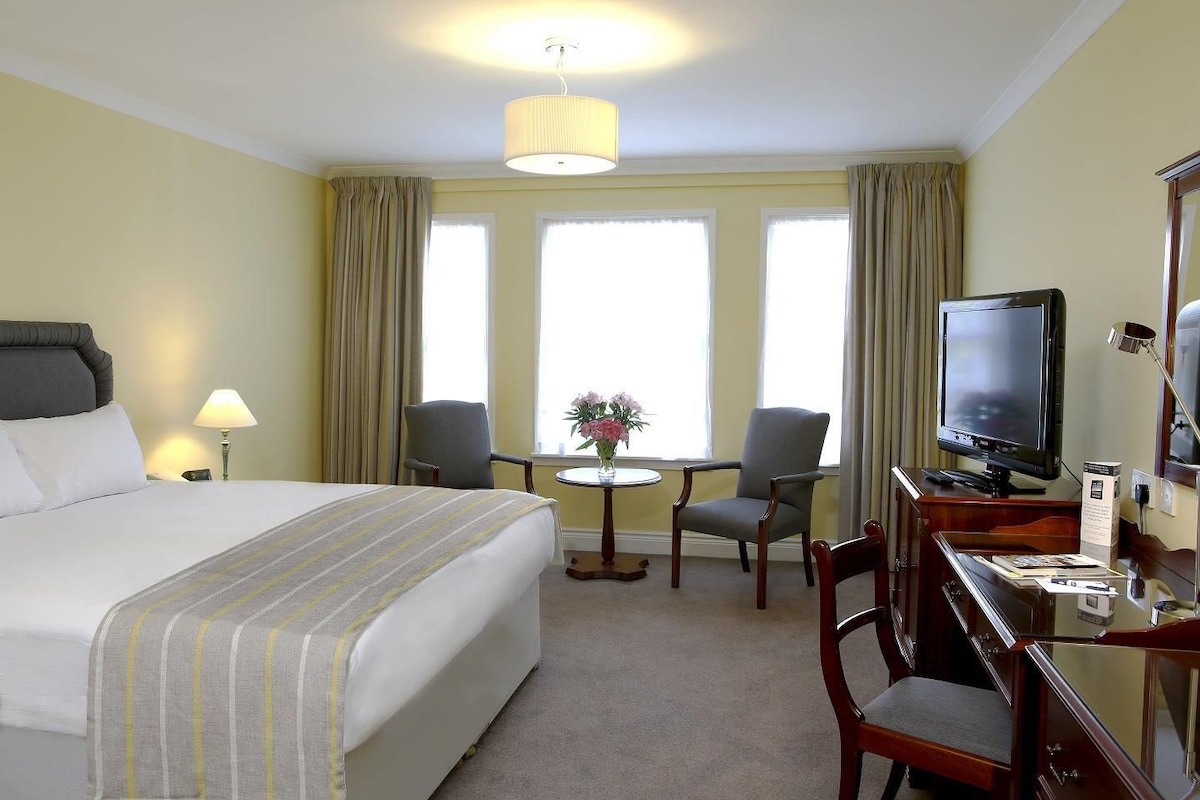 A hotel room with a bed, a desk with a television, two chairs, a small table with flowers, and a lamp.
