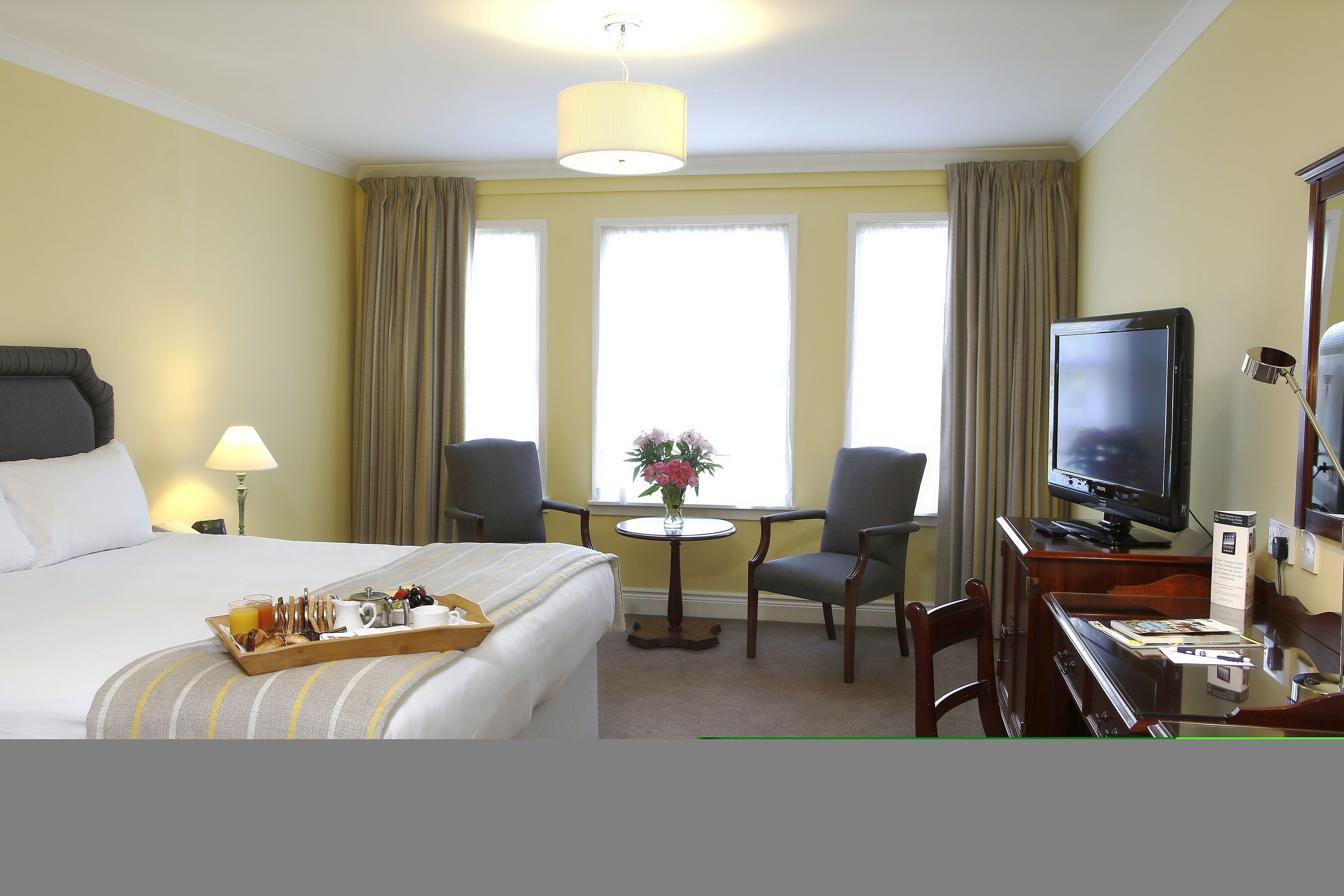 A hotel room with a bed, a desk with a television, a small table with a vase of flowers, and a tray with breakfast items.