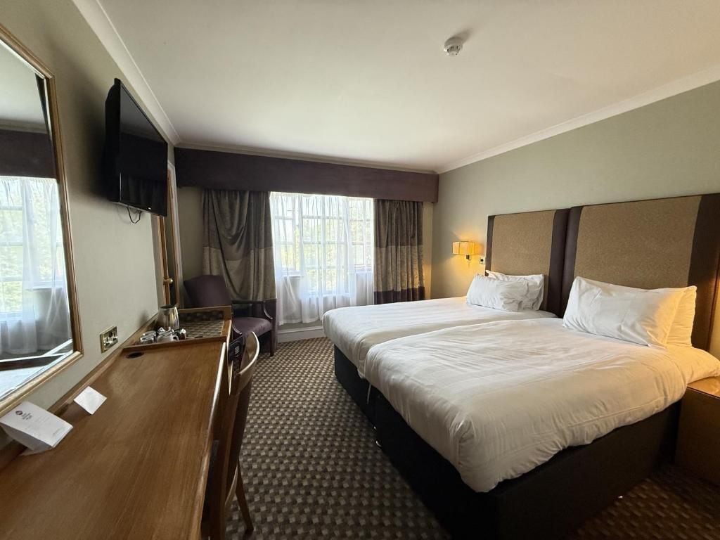 Quarto de hotel com cama, escrivaninha, cadeira, televisão e janela com cortinas.