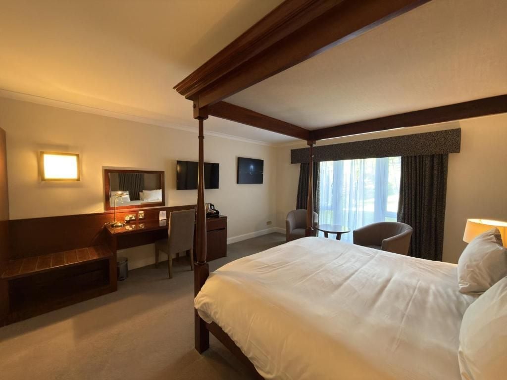 A hotel room with a four-poster bed, a desk with a chair, a small table, and a window with curtains.