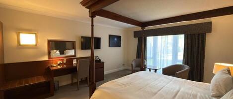 A hotel room with a four-poster bed, a desk with a chair, a small table, and a window with curtains.