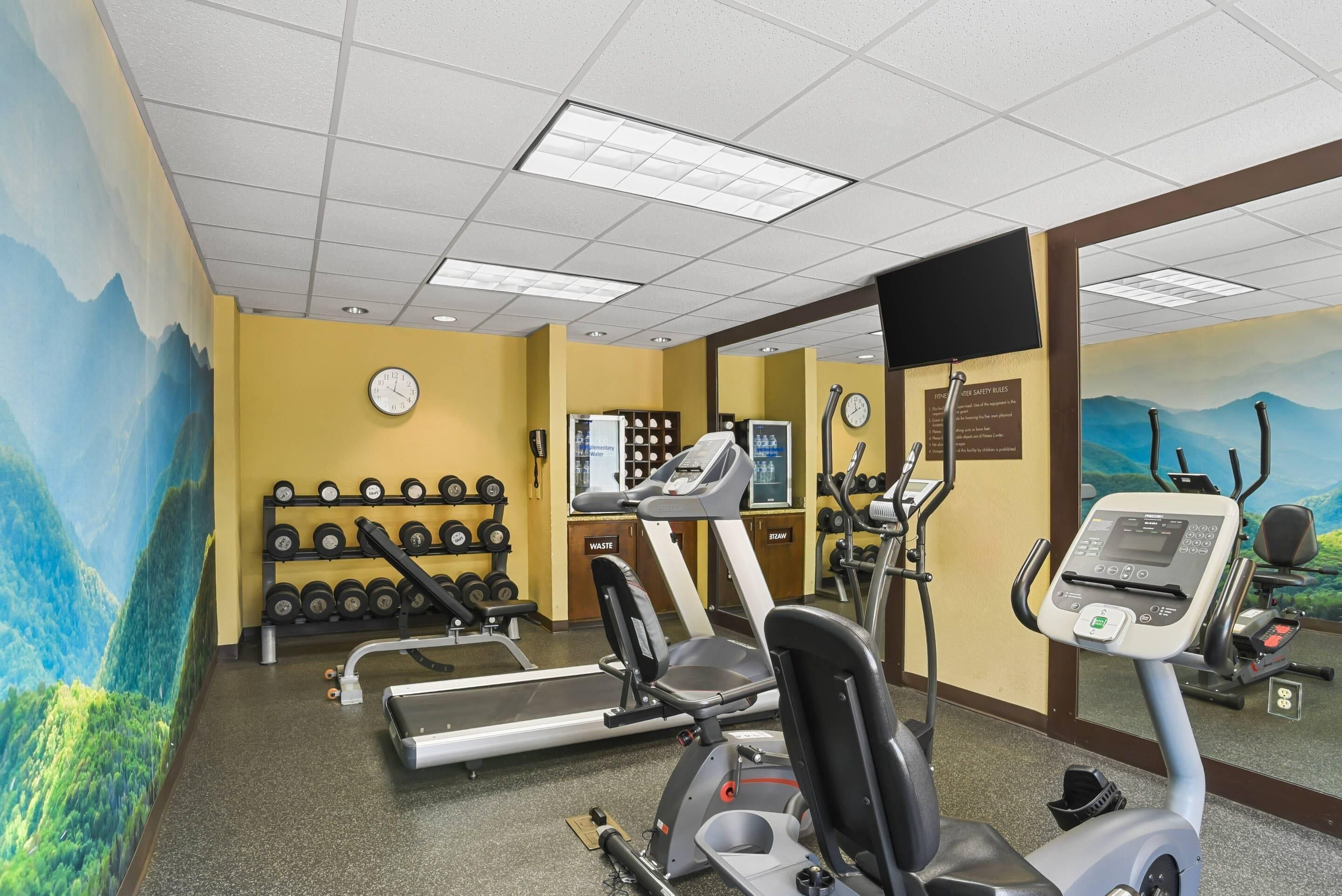 Uma academia com equipamentos de exercício, um relógio de parede e um espelho.