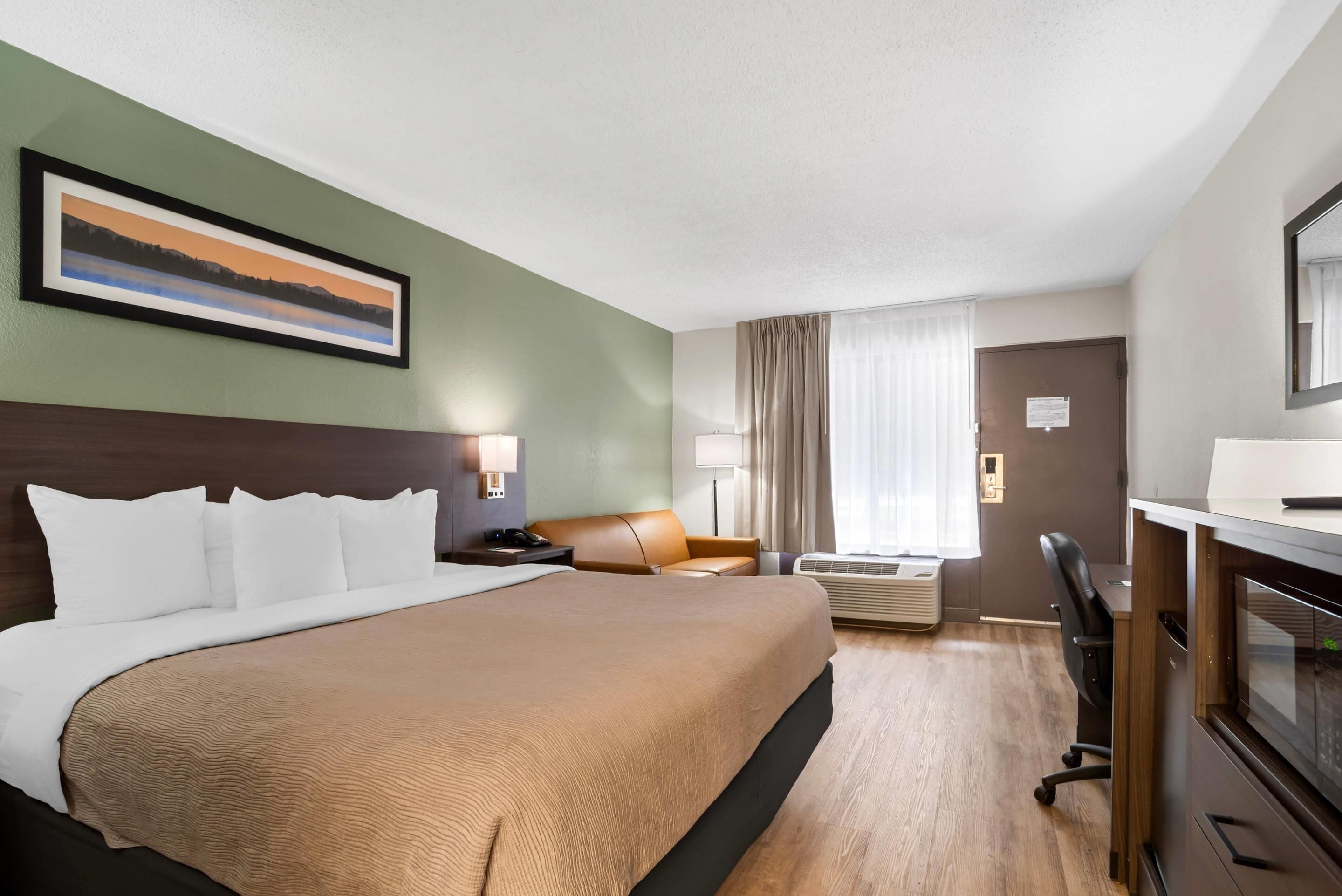 A hotel room with a large bed, a desk, a chair, a lamp, and a painting on the wall.