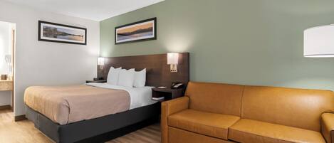 Habitación de hotel con cama, sofá de cuero marrón, mesita de noche con lámpara y dos cuadros en la pared.