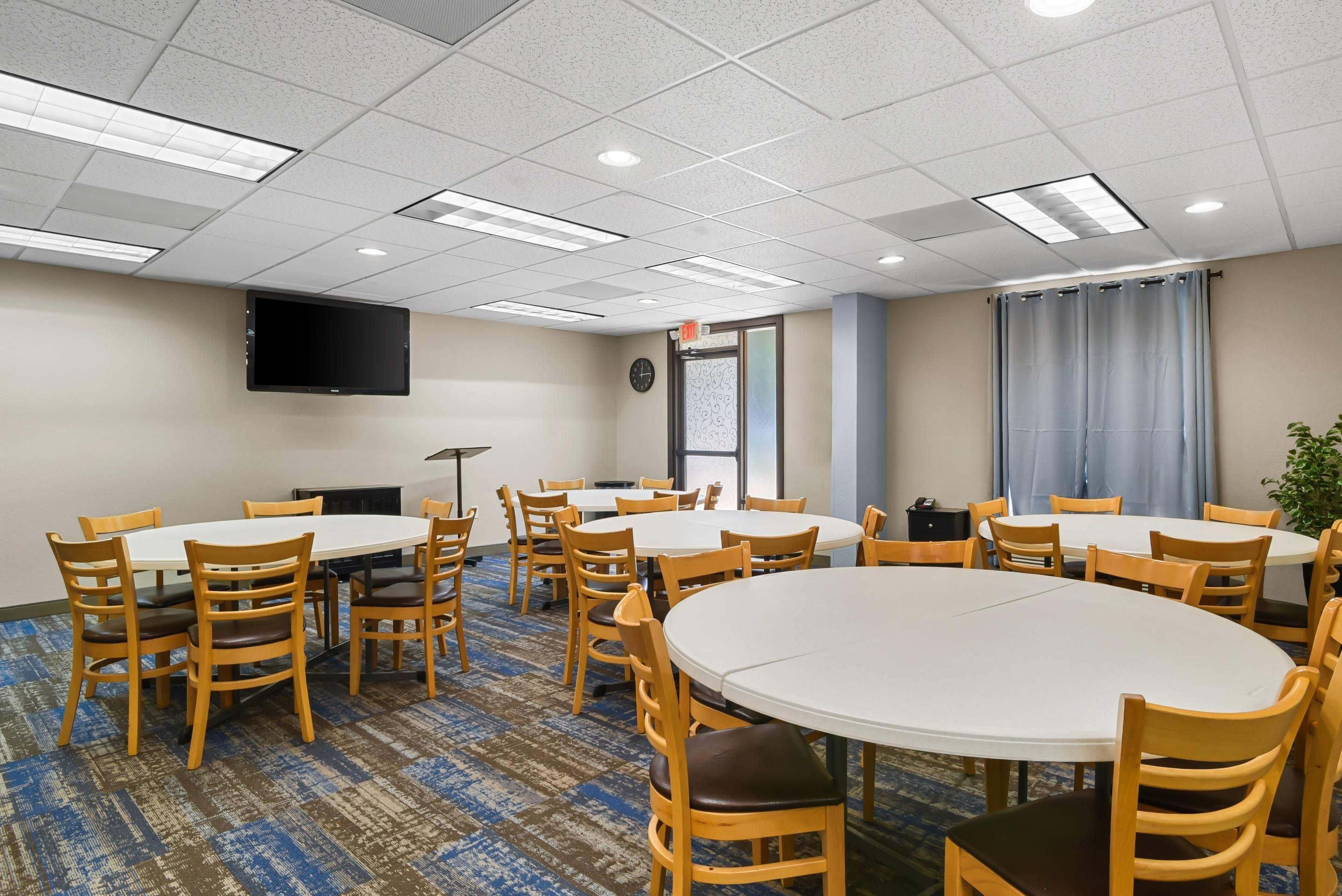 A conference room with multiple round tables and chairs, a flat-screen TV mounted on the wall, and an exit sign above a door.