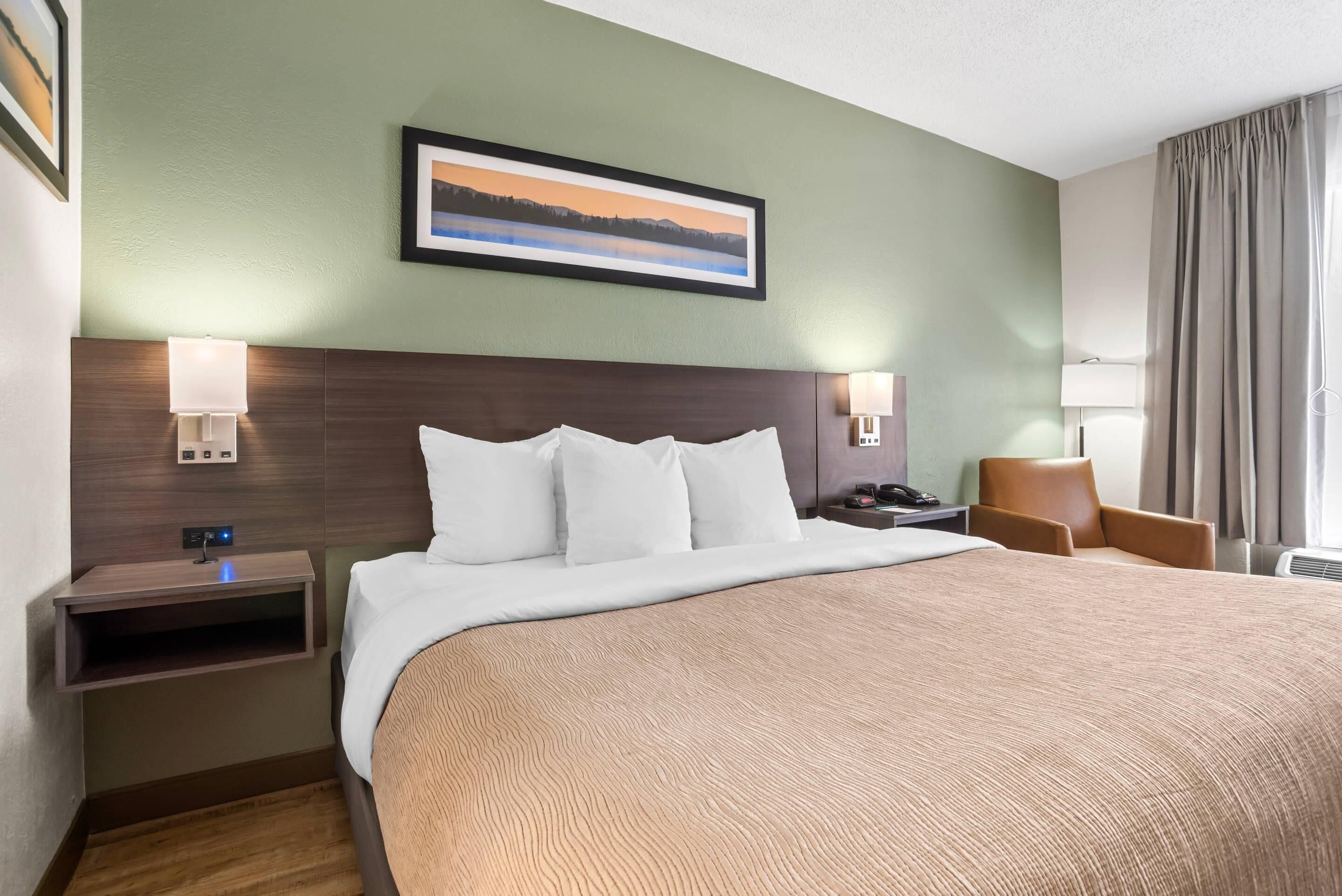 A hotel room with a large bed, a nightstand, a lamp, a chair, and a framed picture on the wall.