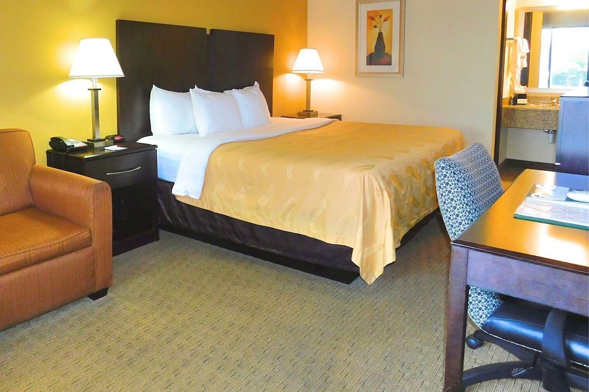A hotel room with a bed, two bedside lamps, a chair, a desk, and a painting on the wall.