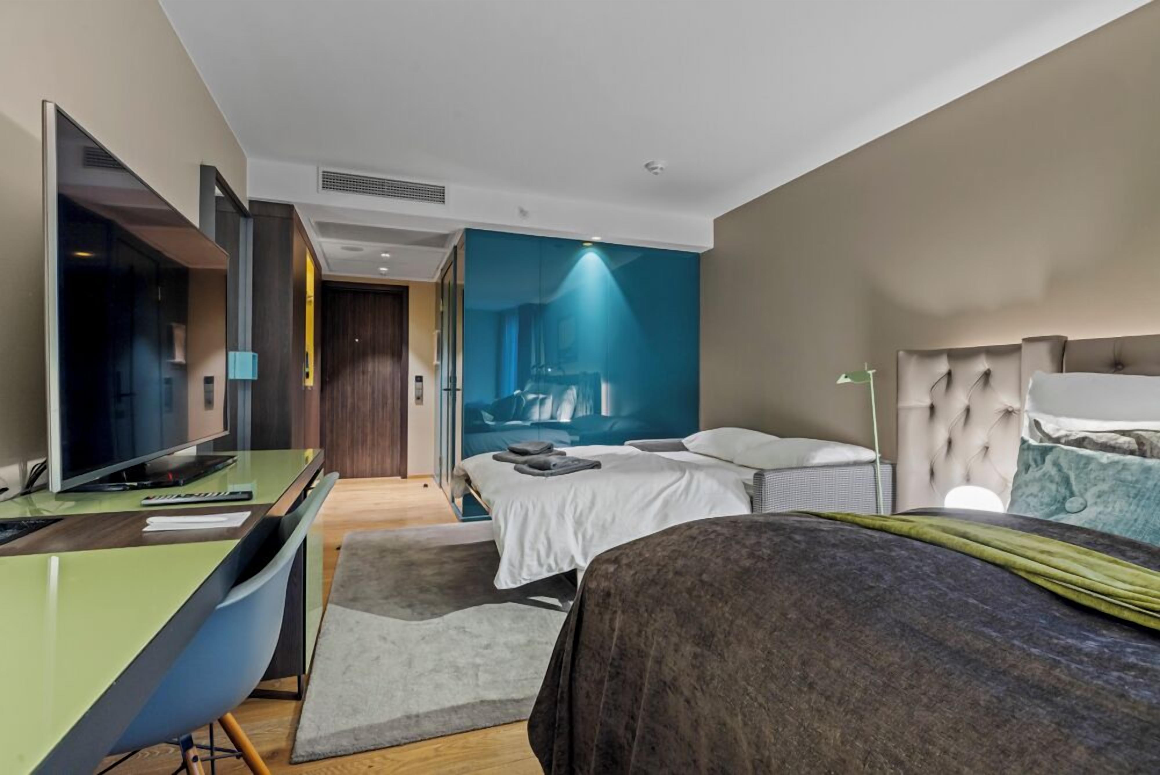 A modern hotel room with a bed, a desk with a computer, a television, and a large window.