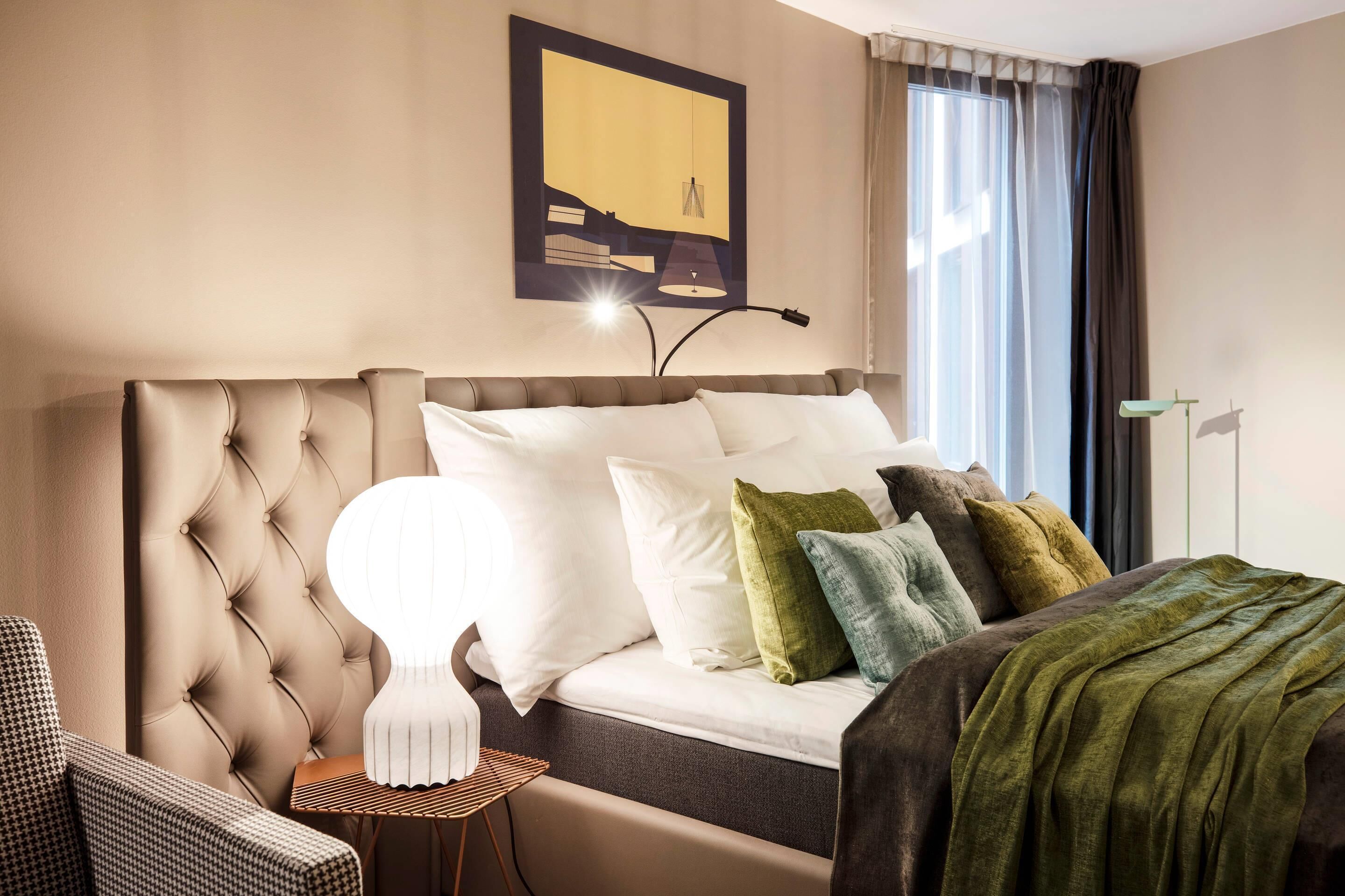 Una camera d'albergo con testiera trapuntata, un letto con molti cuscini, un comodino, una lampada, un quadro appeso alla parete e una finestra con tende.
