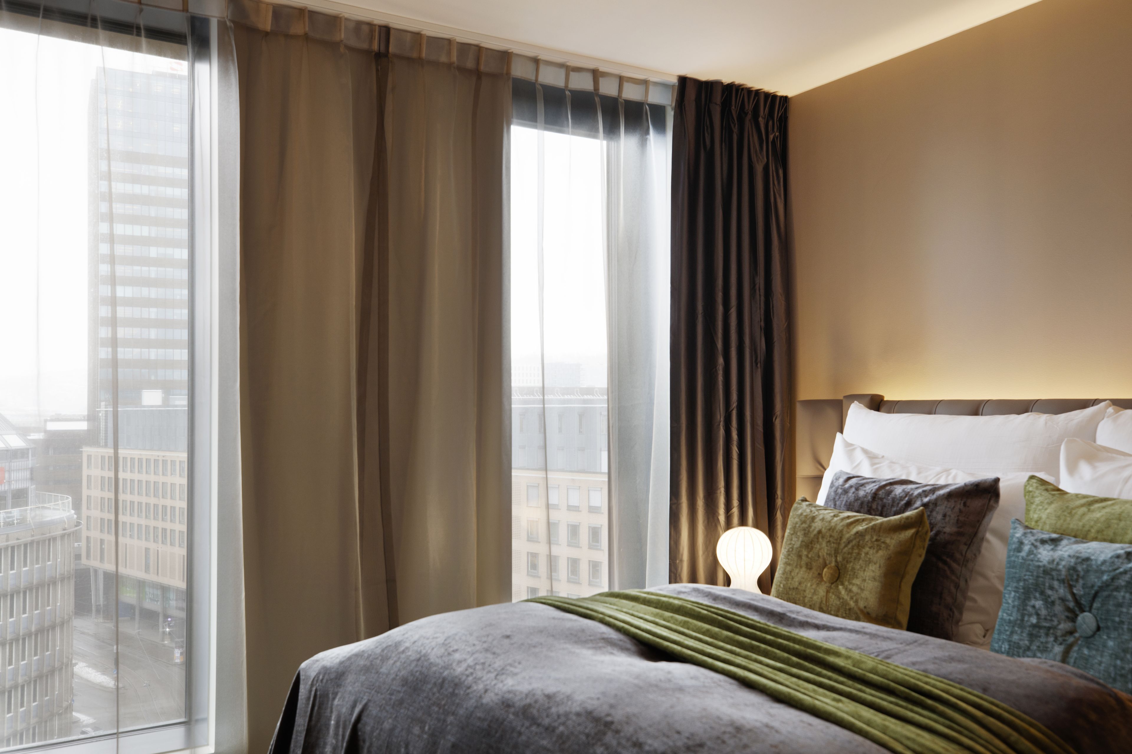 A hotel room with a large window, a bed with multiple pillows, and a nightstand with a lamp.