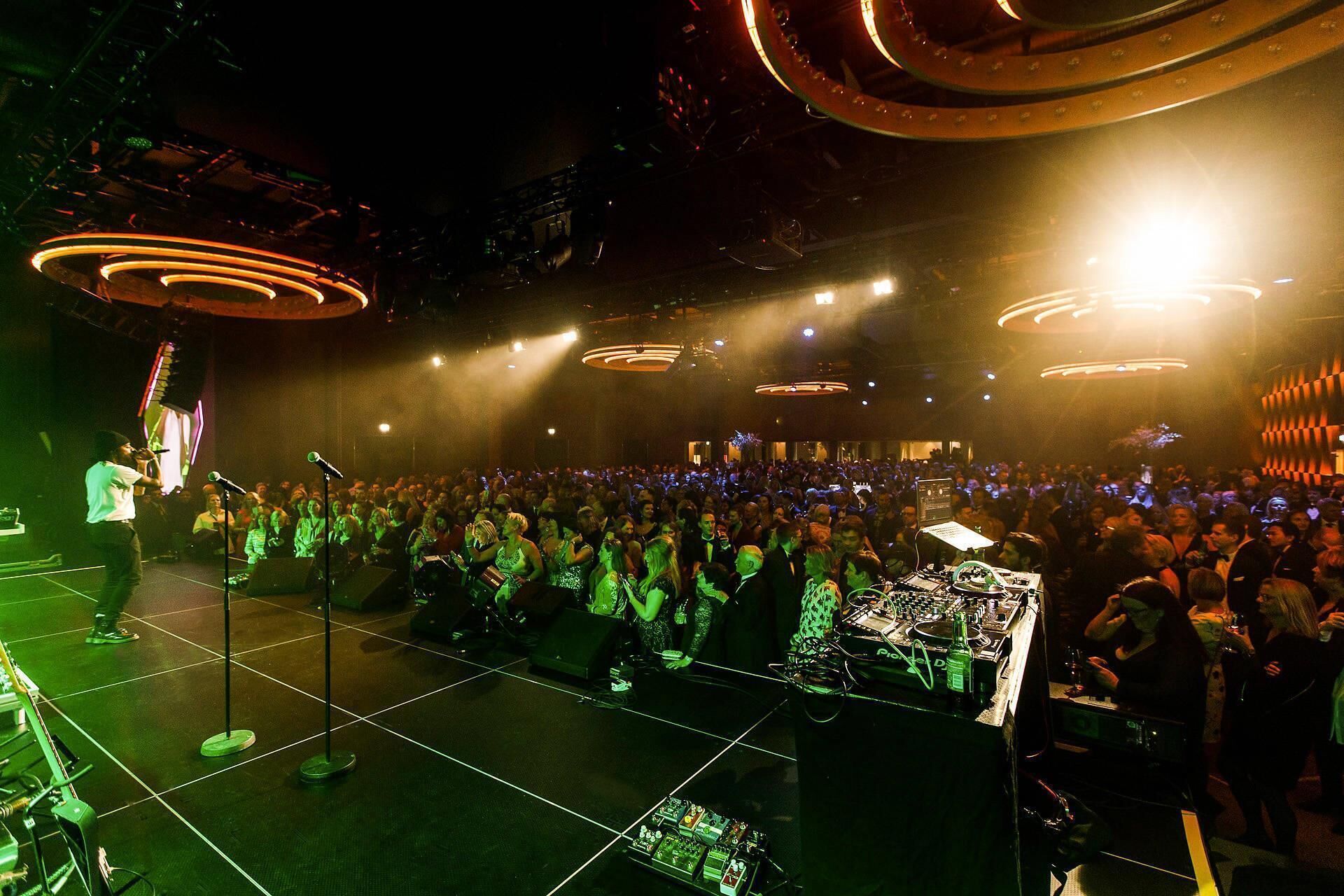 A crowded indoor event with a stage, lighting fixtures, and a DJ setup.