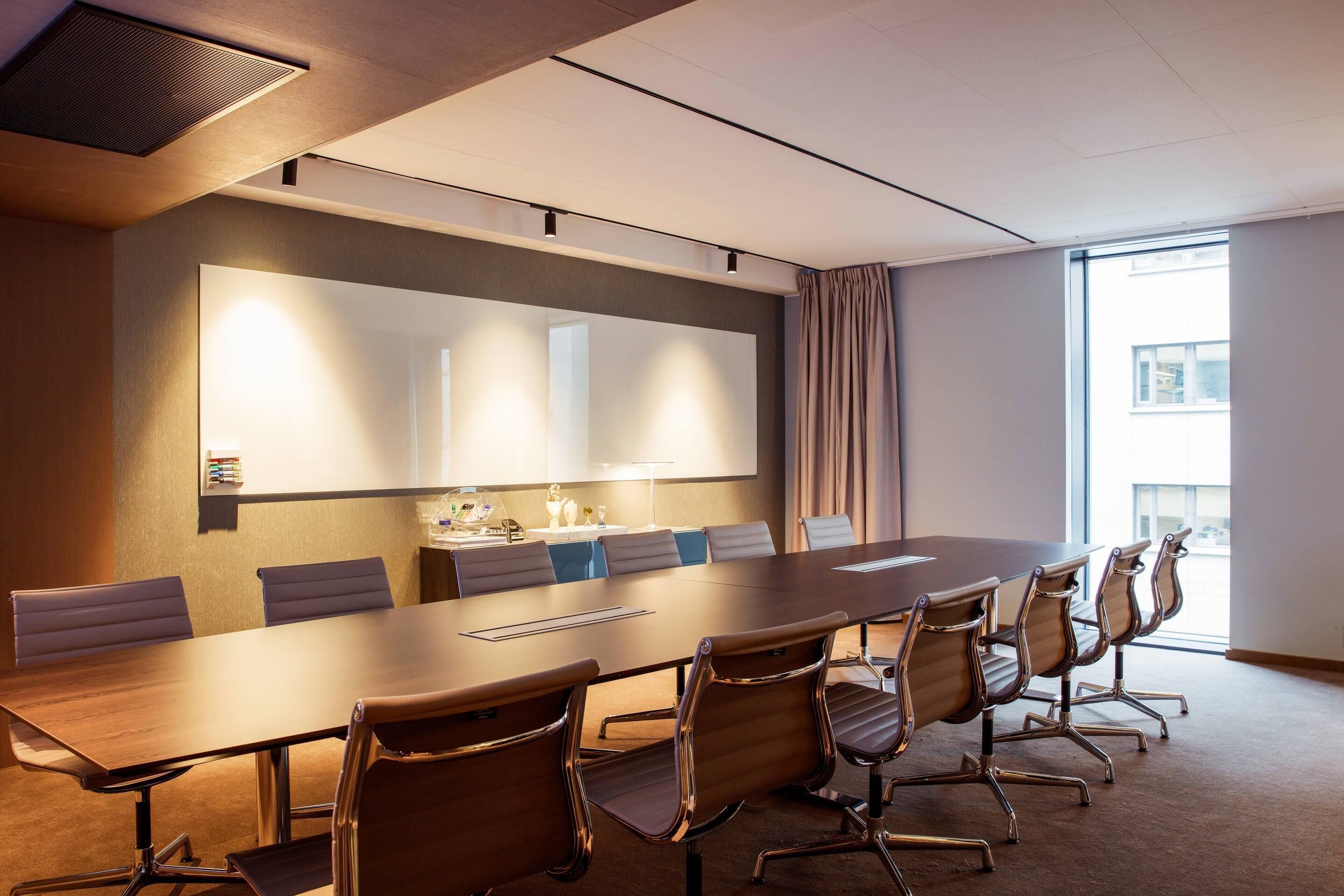 A conference room with a long table, chairs, a whiteboard, and a window with curtains.